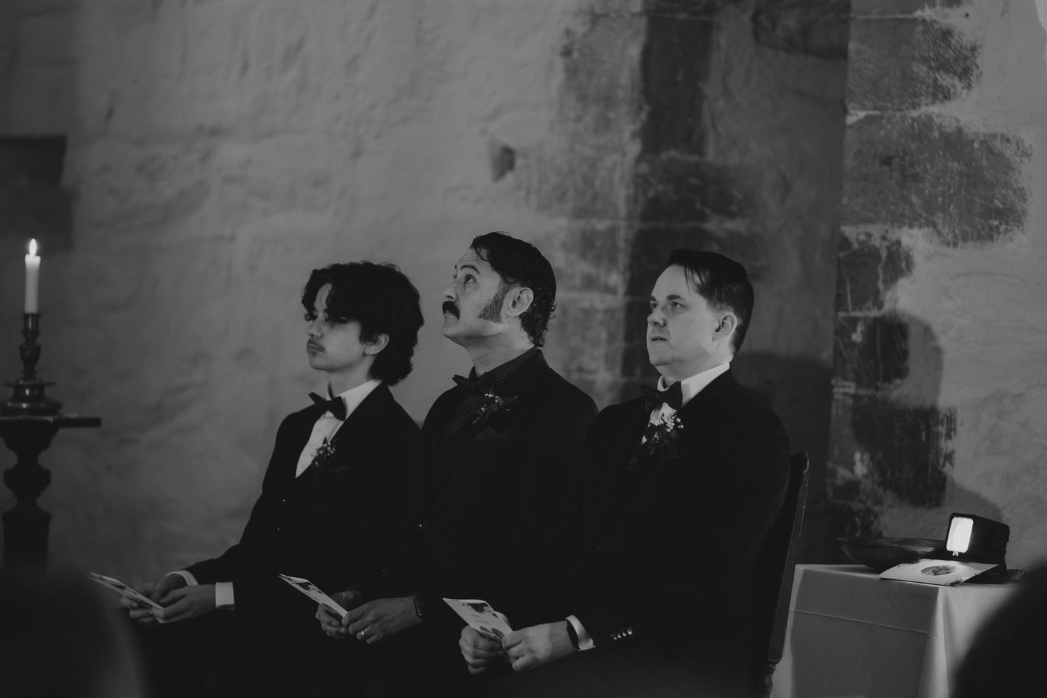 Three groomsmen in black suits sit in a row during a wedding ceremony in a rustic stone chapel.