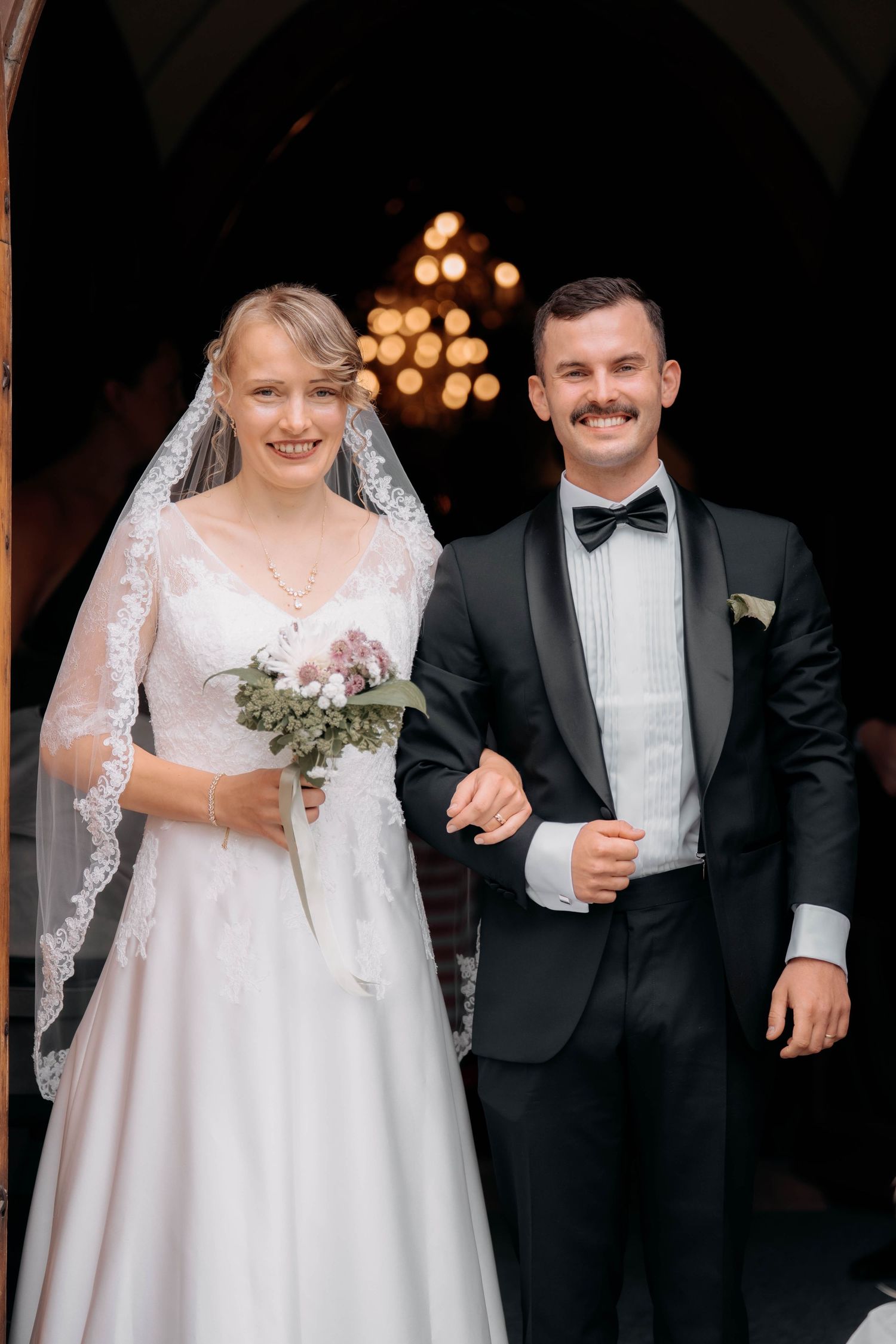 Smiling bride in white lace gown and veil holds bouquet while groom in black tuxedo and bow tie escort her from church.