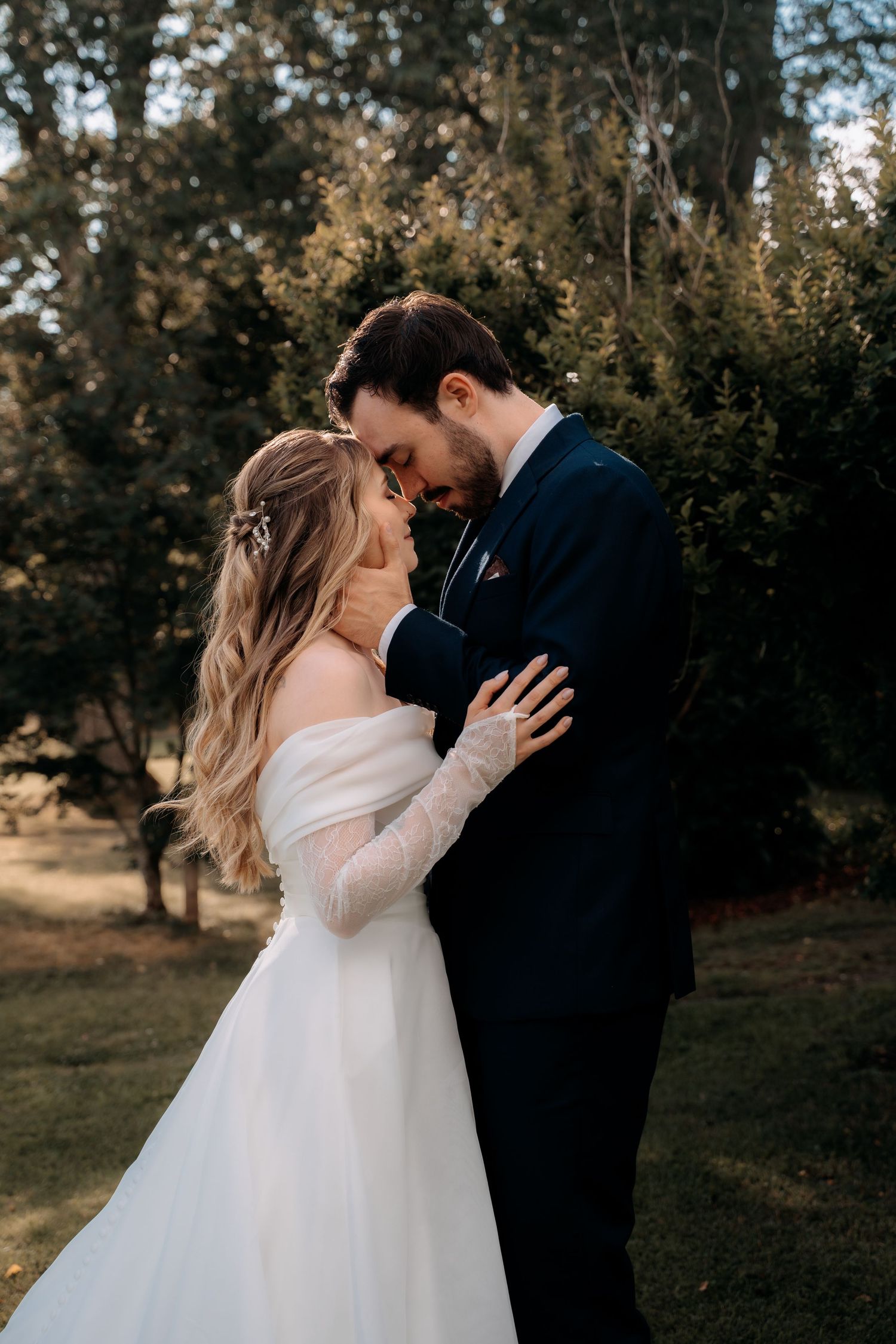 Bride and groom embracing forehead-to-forehead outdoors, bride in off-shoulder gown with long lace sleeves.