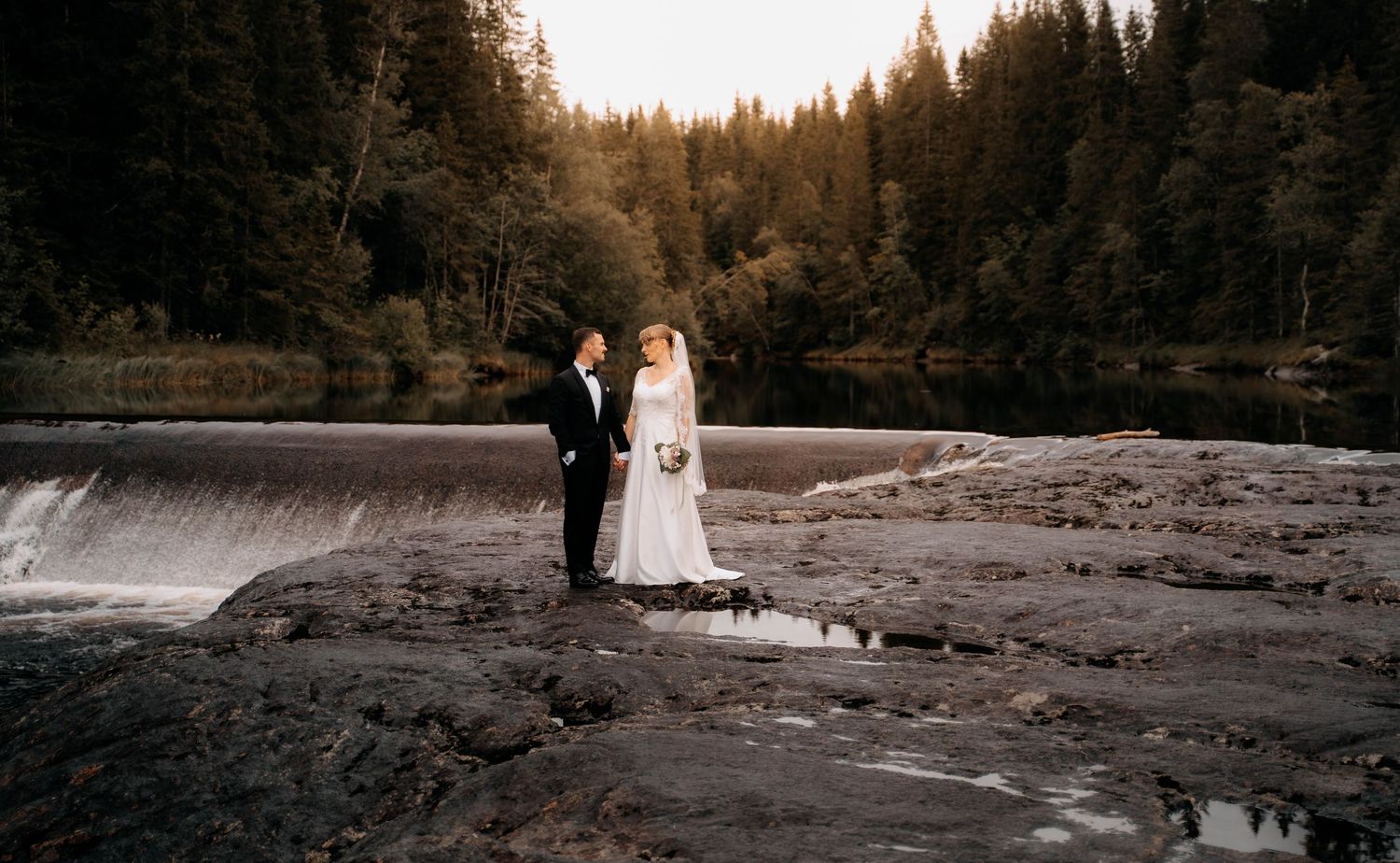 Bride and groom stand on rocky riverbank beside waterfall surrounded by lush evergreen forest at golden hour.