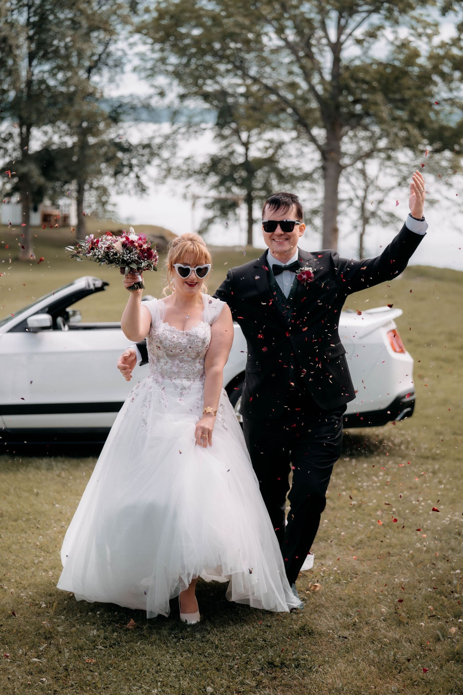 Joyful newlyweds celebrate with confetti wearing sunglasses beside a white convertible car outdoors.