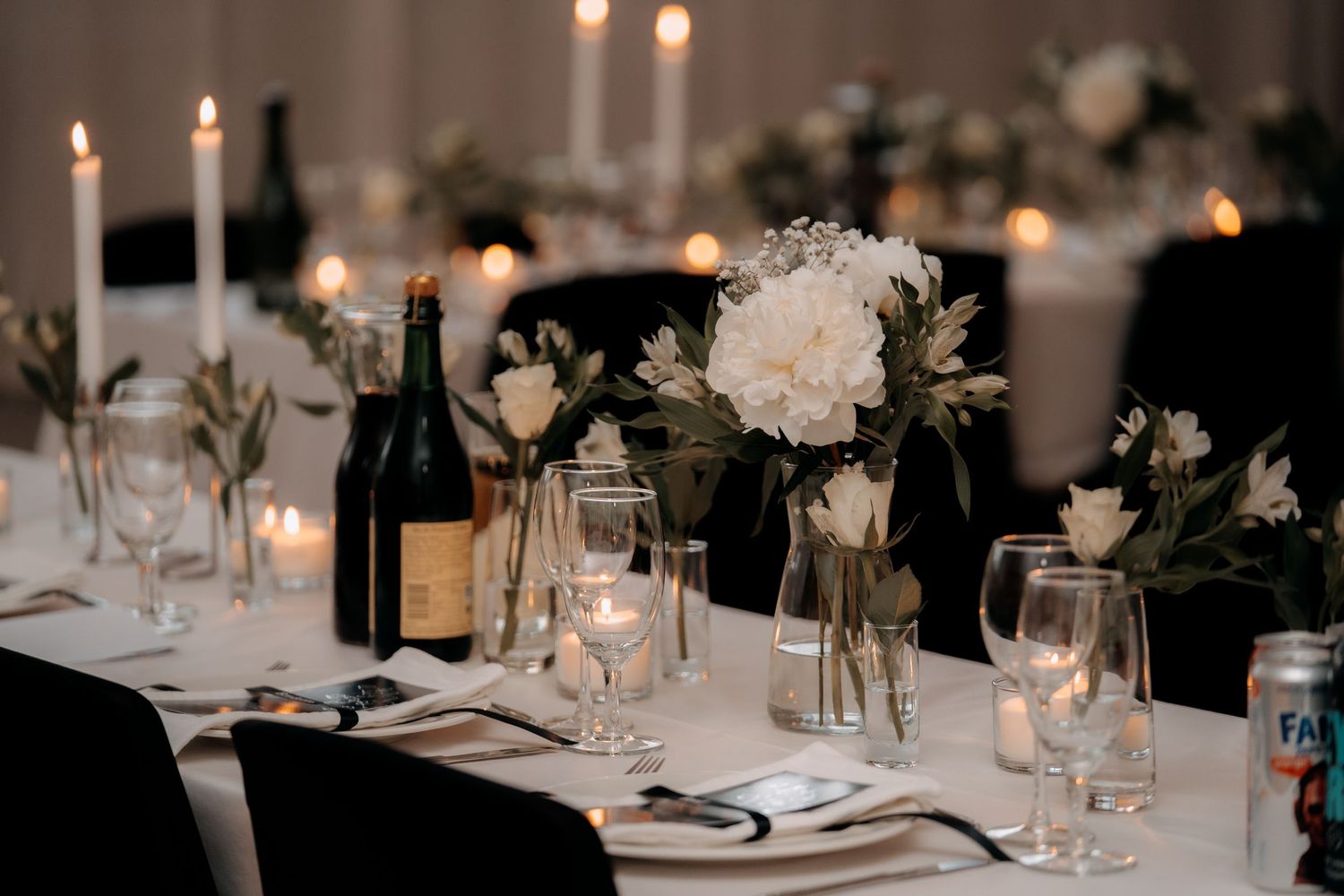 Elegant wedding reception table with white floral centerpieces, taper candles, glassware, and a wine bottle on a white tablecloth.