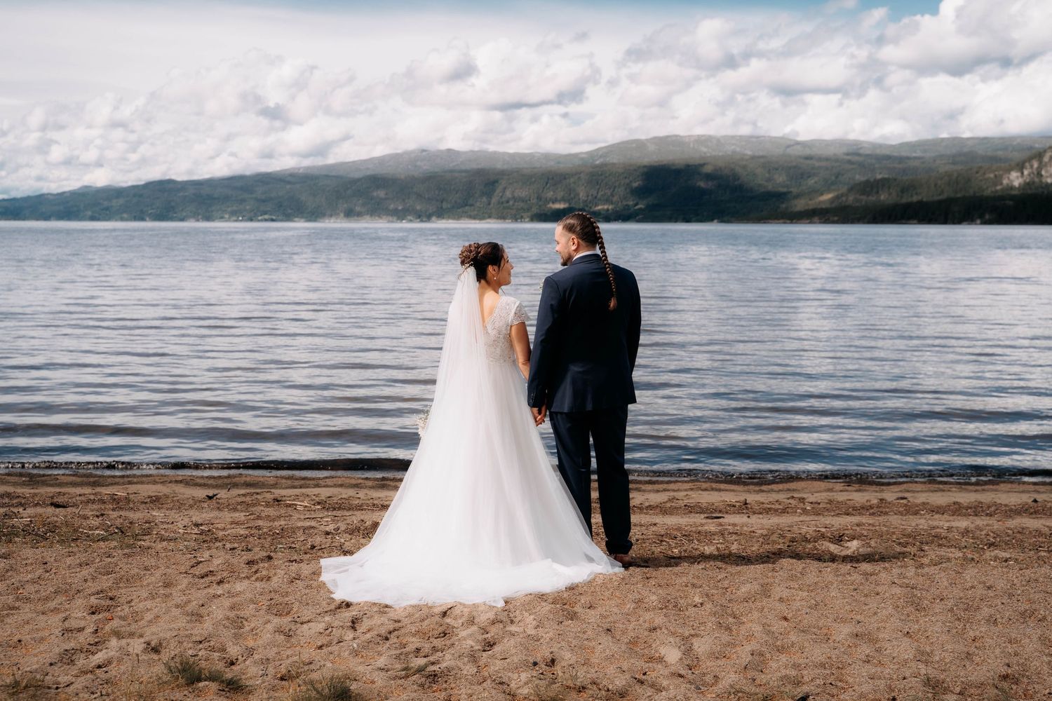 A bride and groom stand hand in hand on a sandy lakeshore, with mountains and cloudy skies in the background.