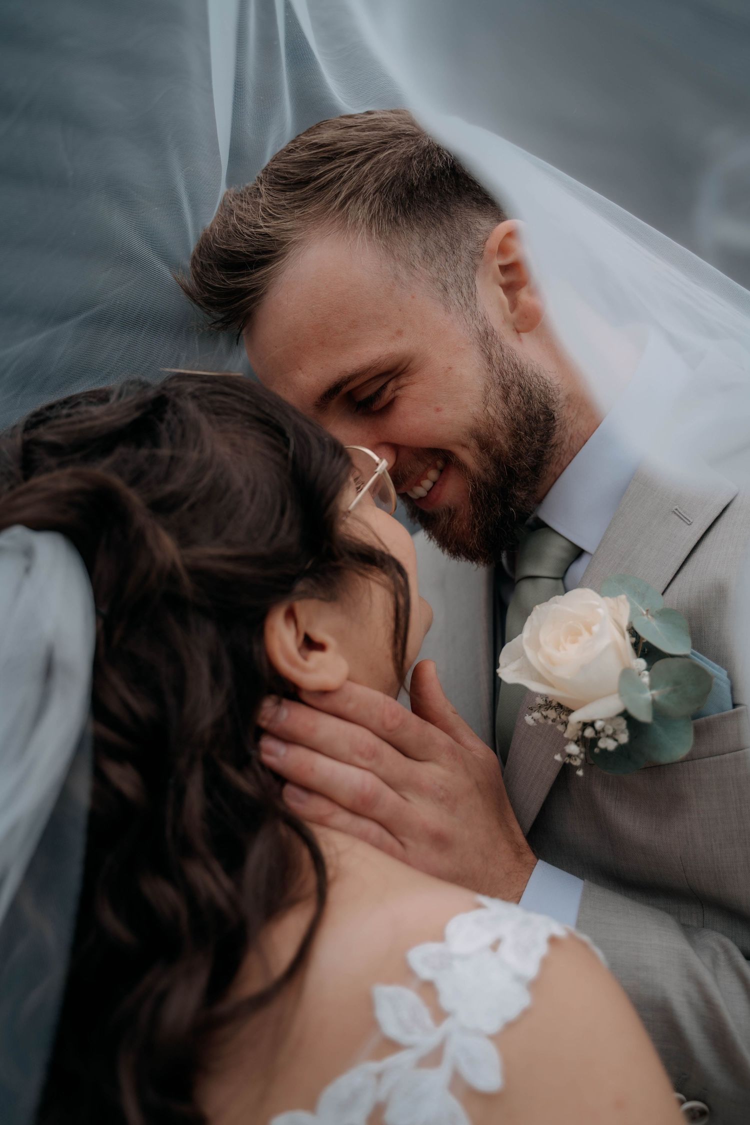 A smiling groom in a gray suit with a rose boutonniere leans close to his bride under a flowing veil.