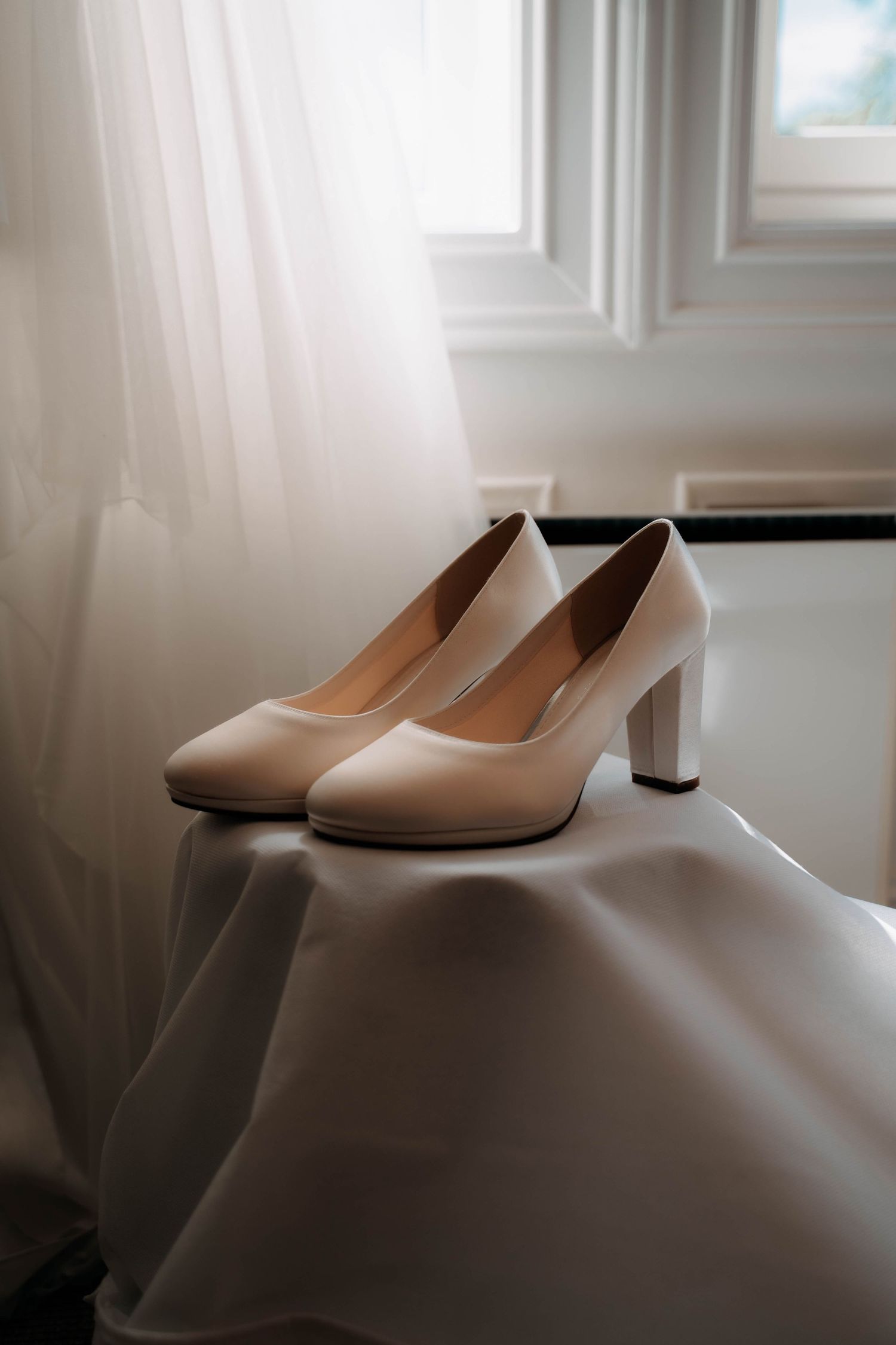 Elegant ivory block heel bridal shoes displayed on white fabric near a sunlit window.