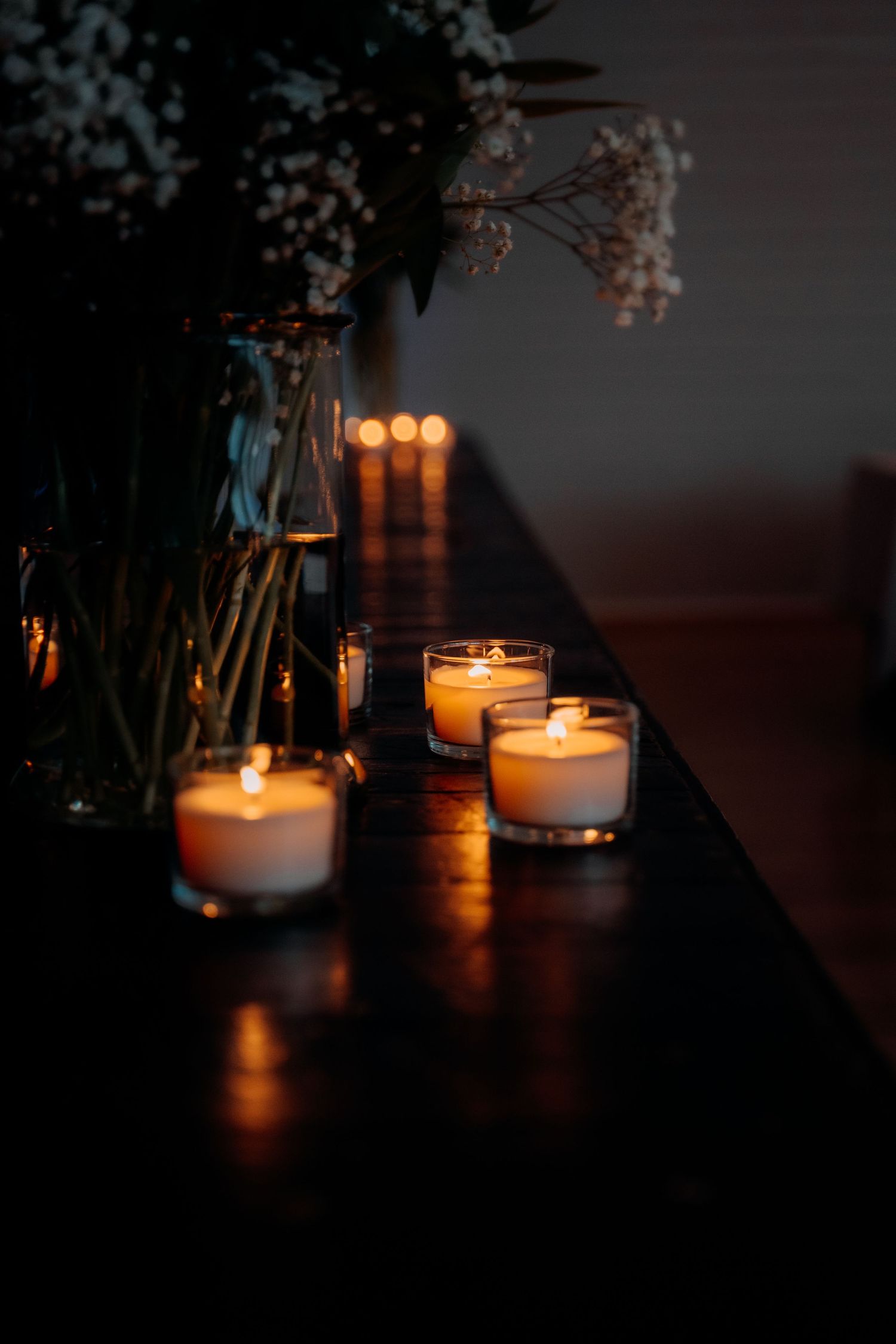 Romantic wedding reception table decorated with glowing votive candles and delicate floral arrangements in dim lighting.