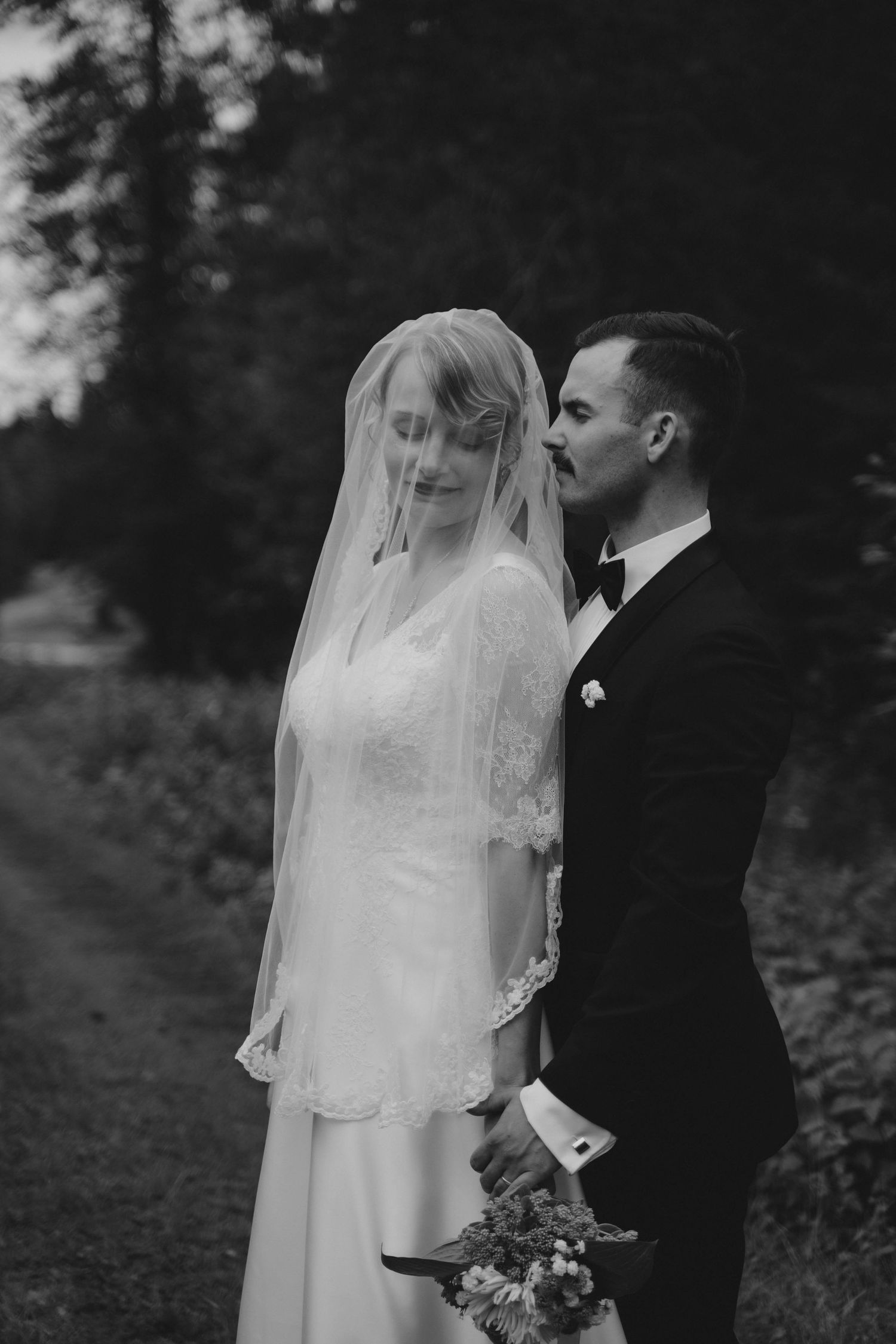 Black and white wedding photo of bride in veil and groom in tuxedo standing together outdoors among trees.