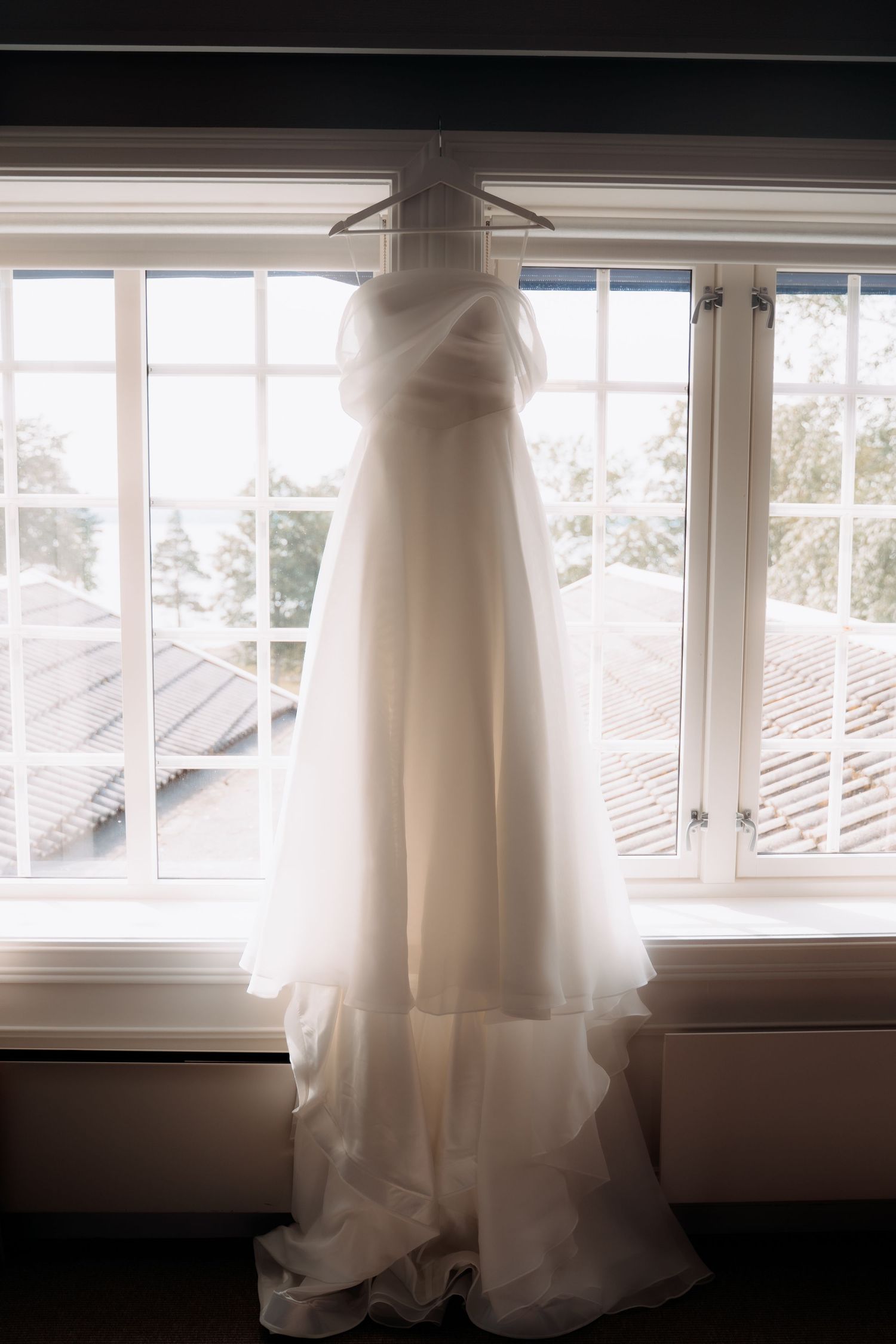White wedding dress hanging on a hanger in front of bright windows with natural light streaming through.