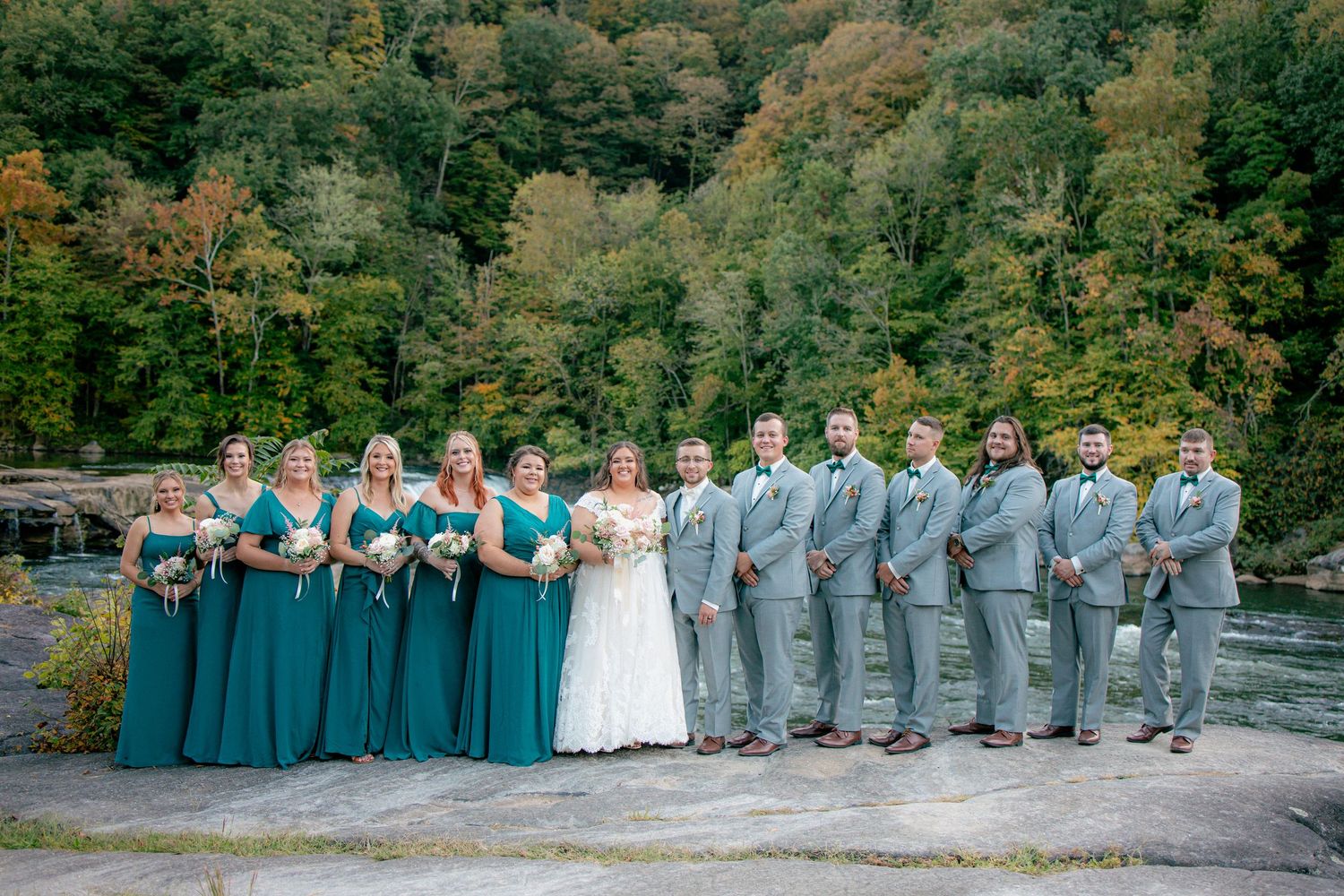Bride and groom pose with wedding party in teal dresses and gray suits on rocky riverbank with autumn forest backdrop.