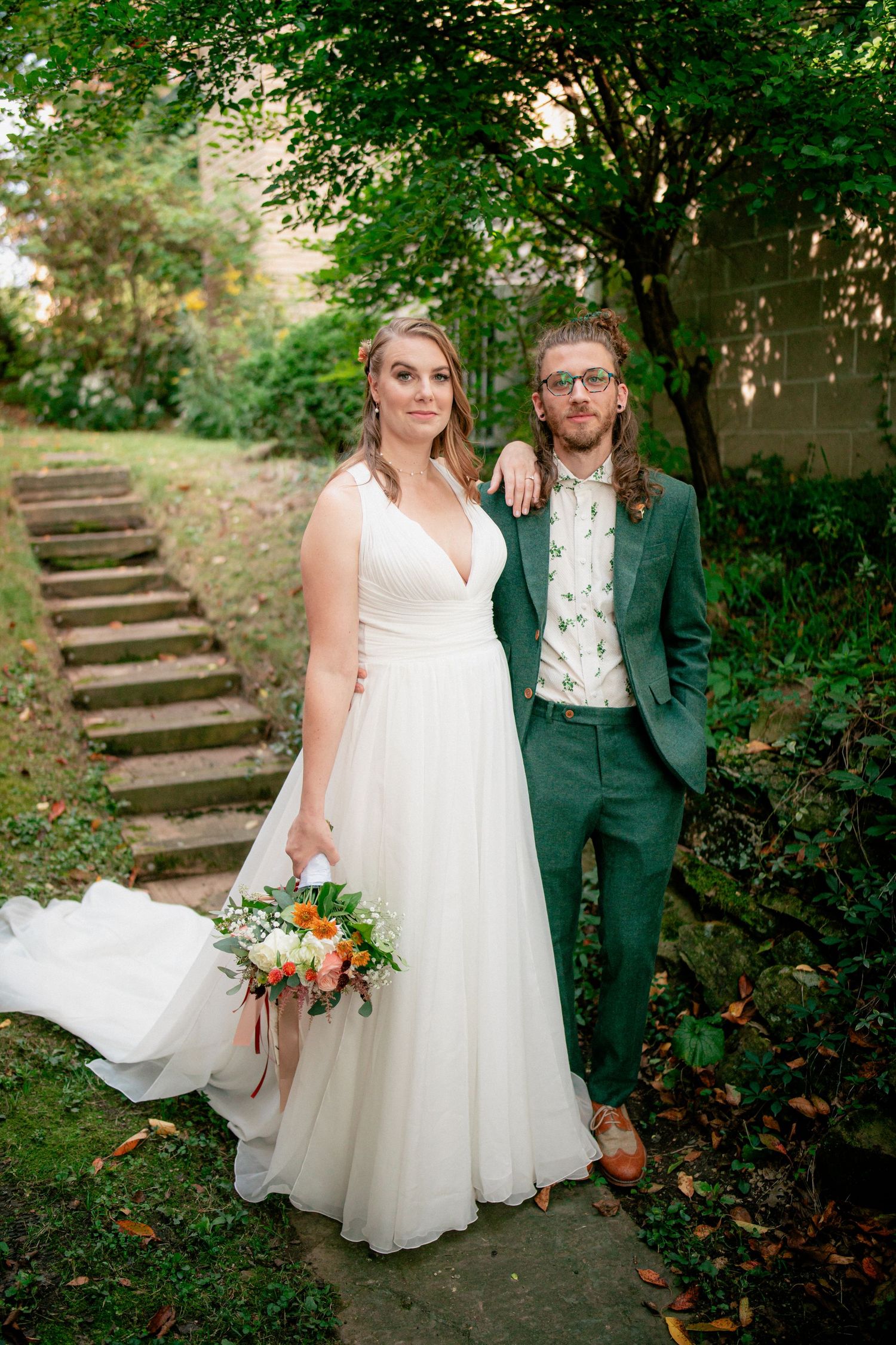 Bride and groom pose together on garden stone path surrounded by lush greenery and stone steps.