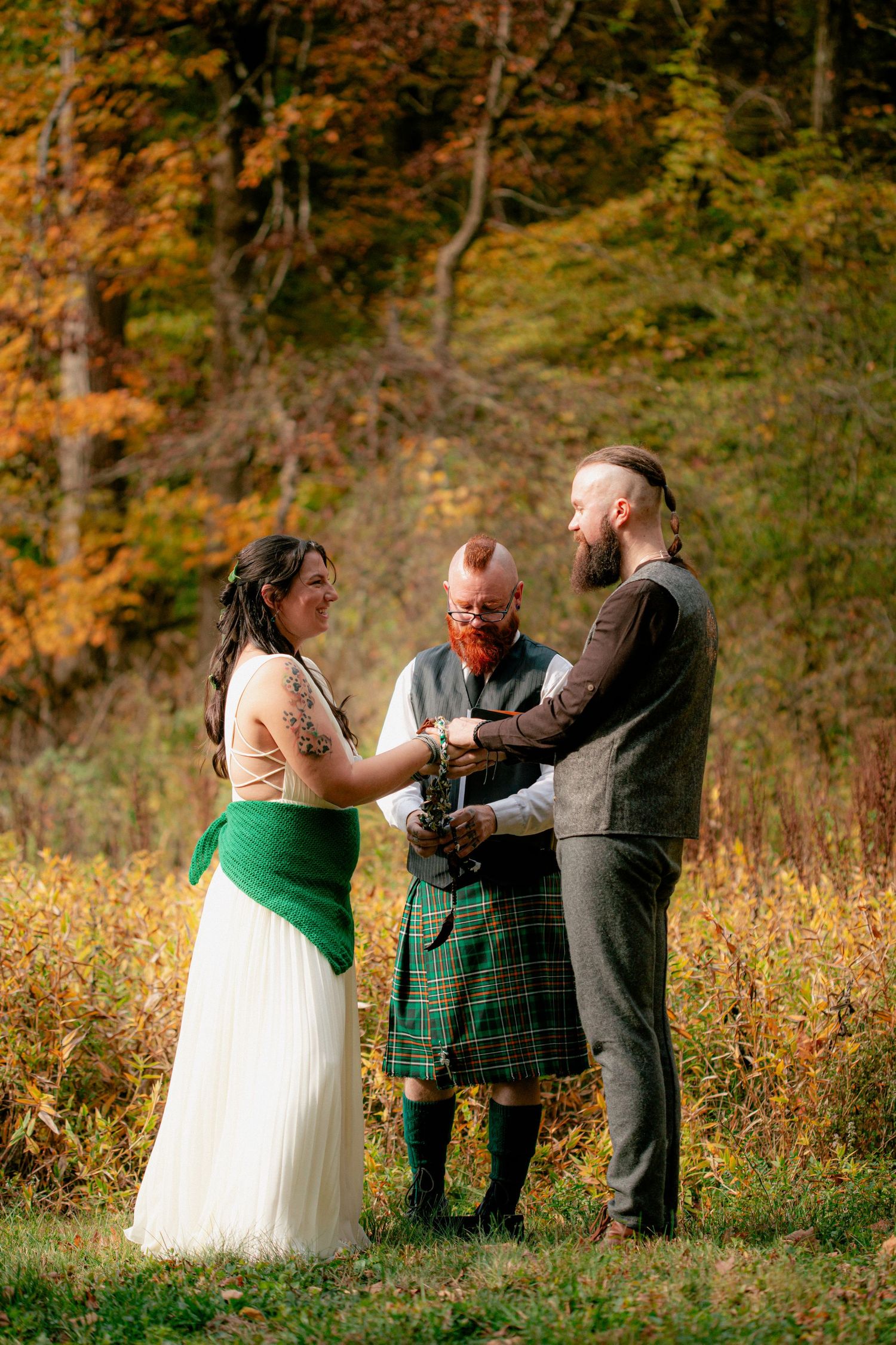 Bride and groom hold hands during outdoor fall wedding ceremony officiated by man in green tartan kilt.