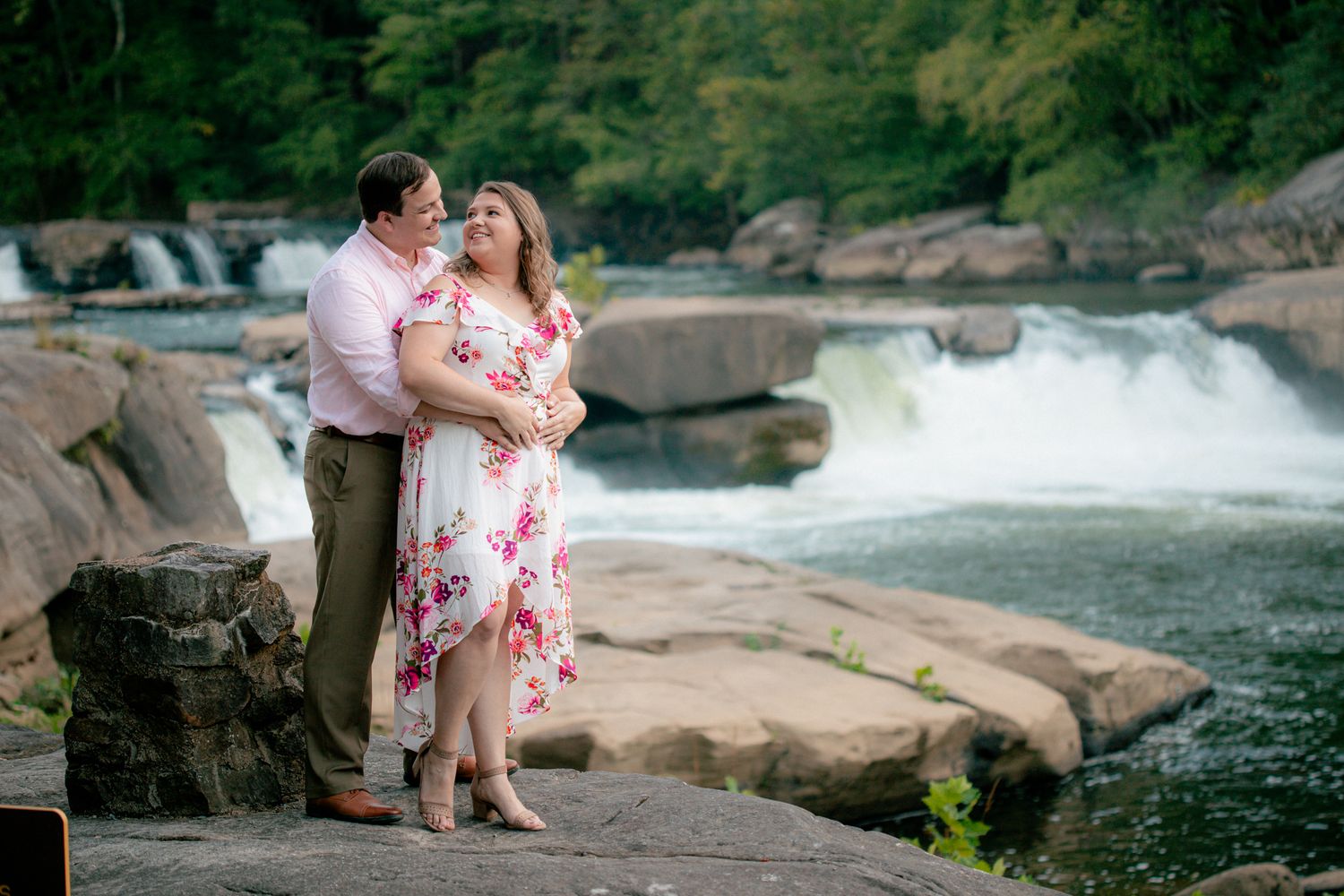 Couple posing on rocks with a rushing waterfall backdrop, woman in floral dress smiling warmly.