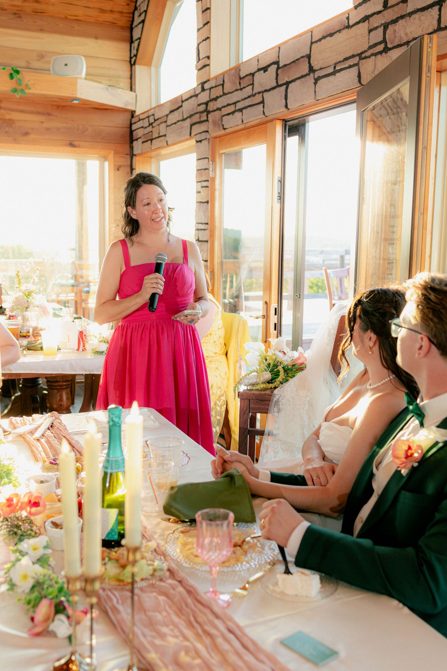 Bridesmaid in pink dress gives wedding toast speech to bride and groom at elegant reception venue.