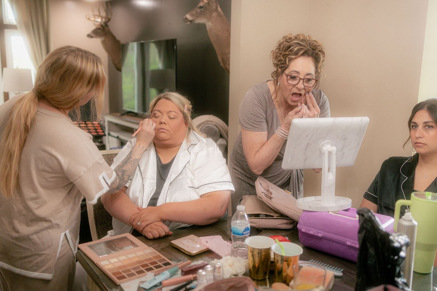 Women getting professional makeup applied at a vanity mirror station with cosmetics and beauty products.