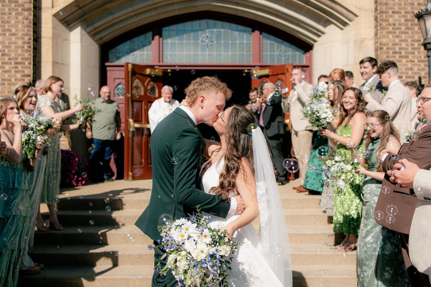 Newlyweds share first kiss on church steps surrounded by cheering guests holding floral bouquets.