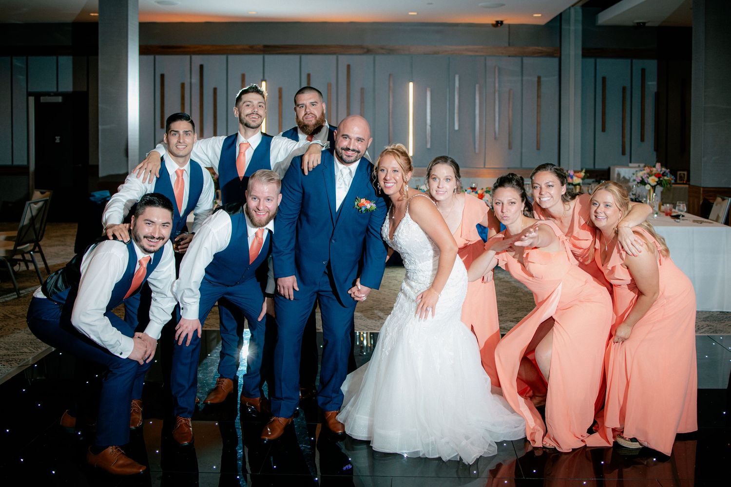 Wedding party posing together on dance floor with groomsmen in navy suits and bridesmaids in coral.