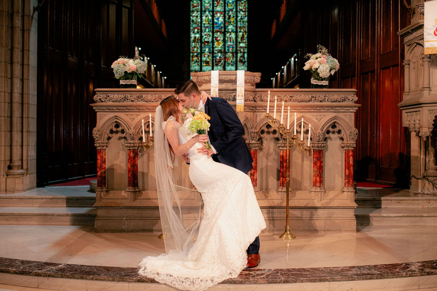 Newlyweds share romantic dip kiss at ornate church altar adorned with candles and white flowers.