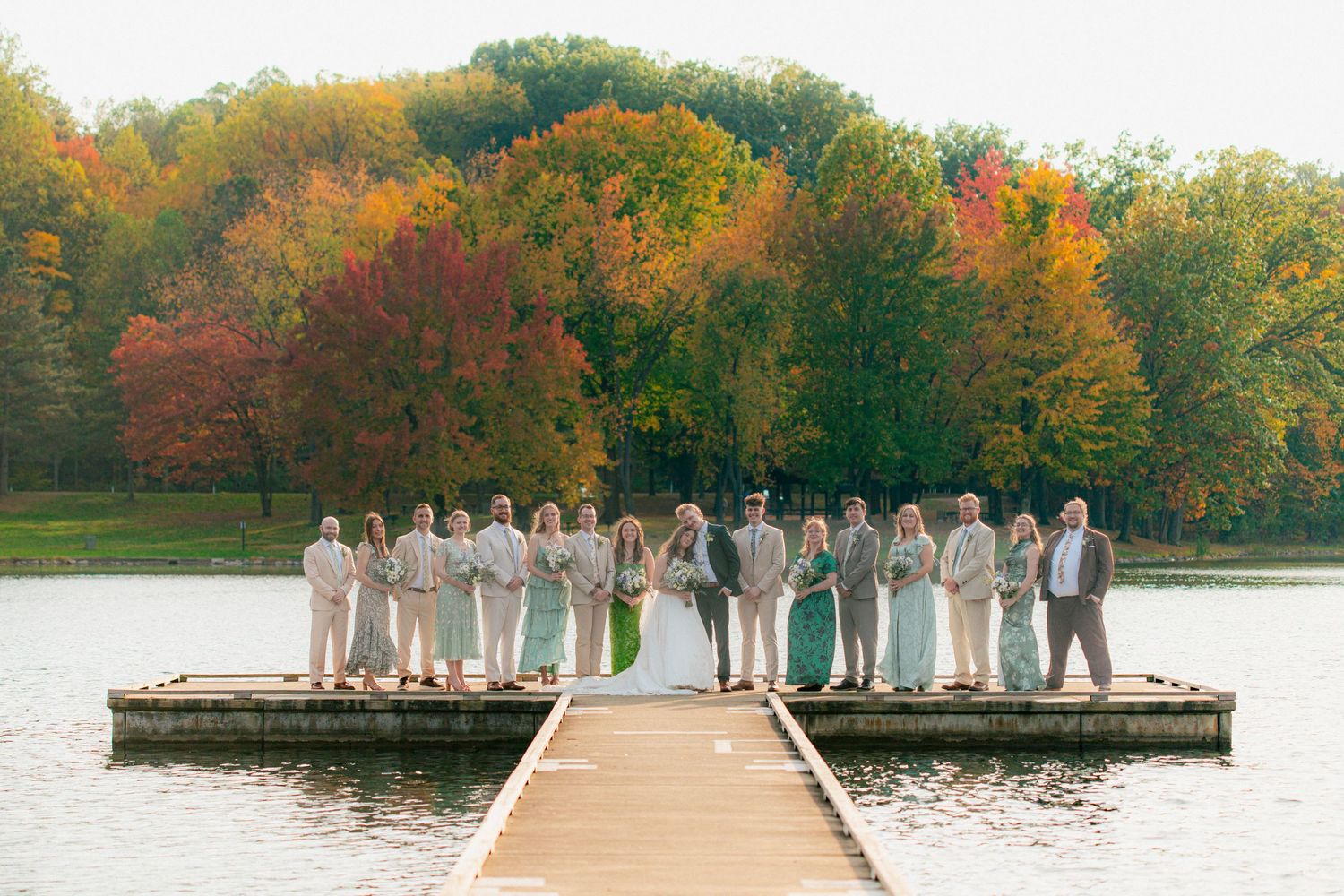 Wedding party posing on a dock surrounded by a lake with vibrant autumn foliage in the background.
