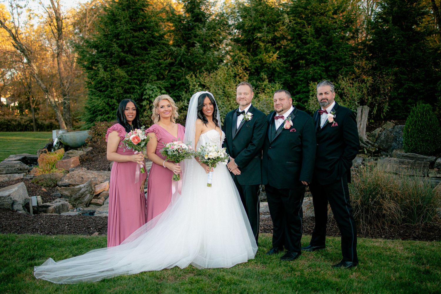 Bride in white ballgown poses with bridesmaids in pink dresses and groomsmen in black tuxedos in an outdoor garden setting.