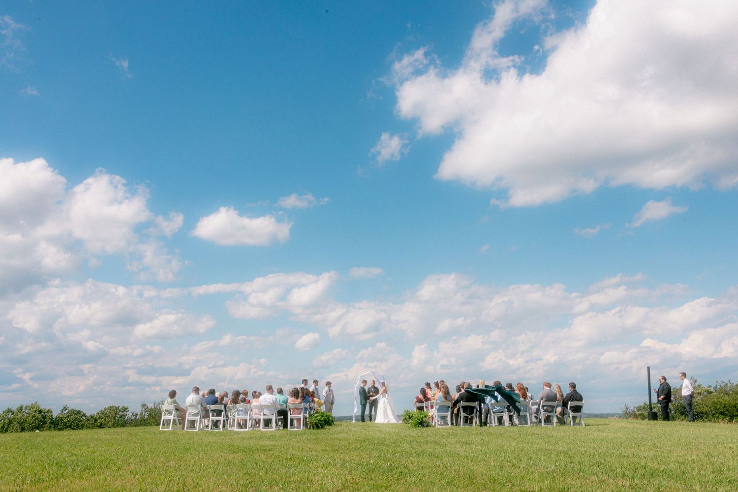 Outdoor wedding ceremony on green lawn under blue sky with white clouds, guests seated in white chairs around couple.