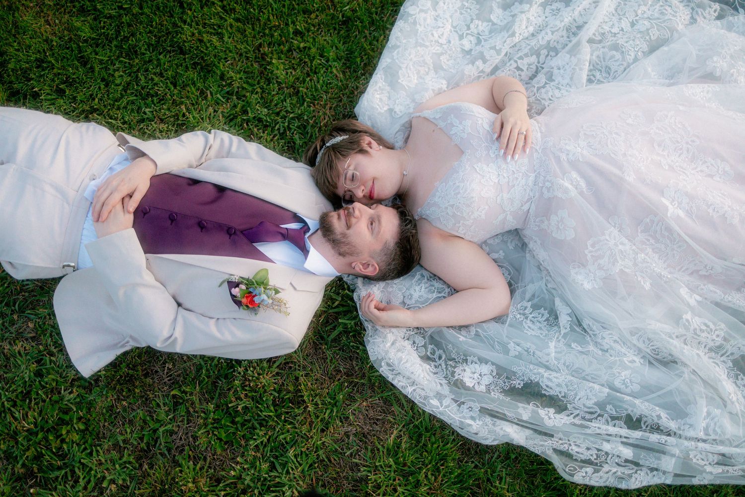 Bride and groom lying on green grass, wearing wedding attire, sharing a romantic moment in an aerial wedding photo.