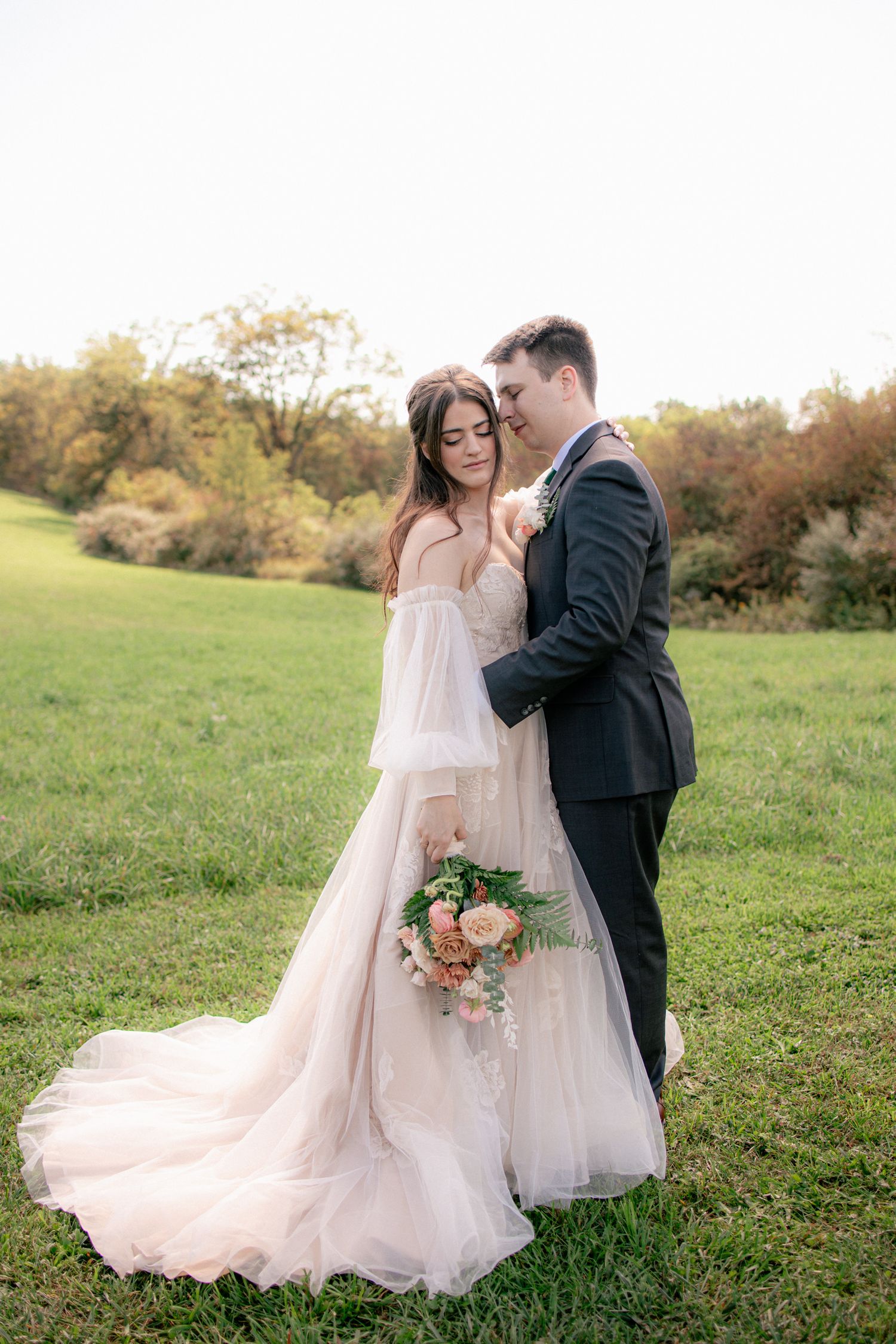 Bride in flowing blush tulle gown with train holds bouquet as groom in dark suit embraces her in a green field.