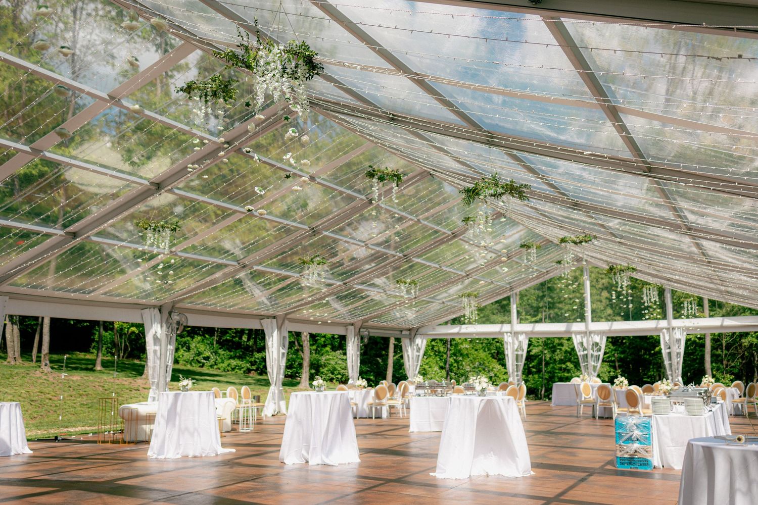 Clear tent wedding reception space with white draped tables, wood floors, and hanging floral decor.