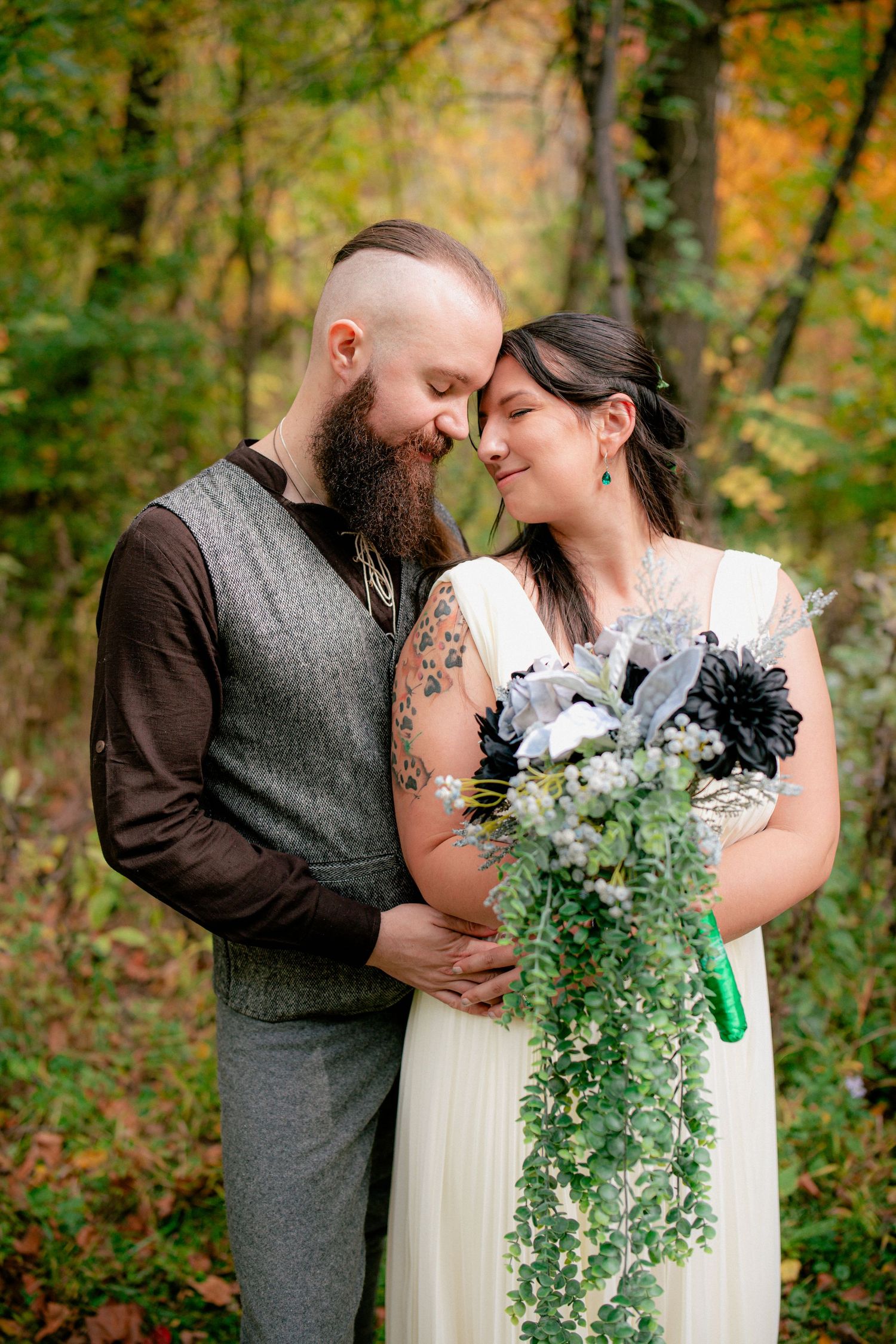 Newlyweds share a tender forehead touch as bride holds cascading green and black bouquet in fall forest.