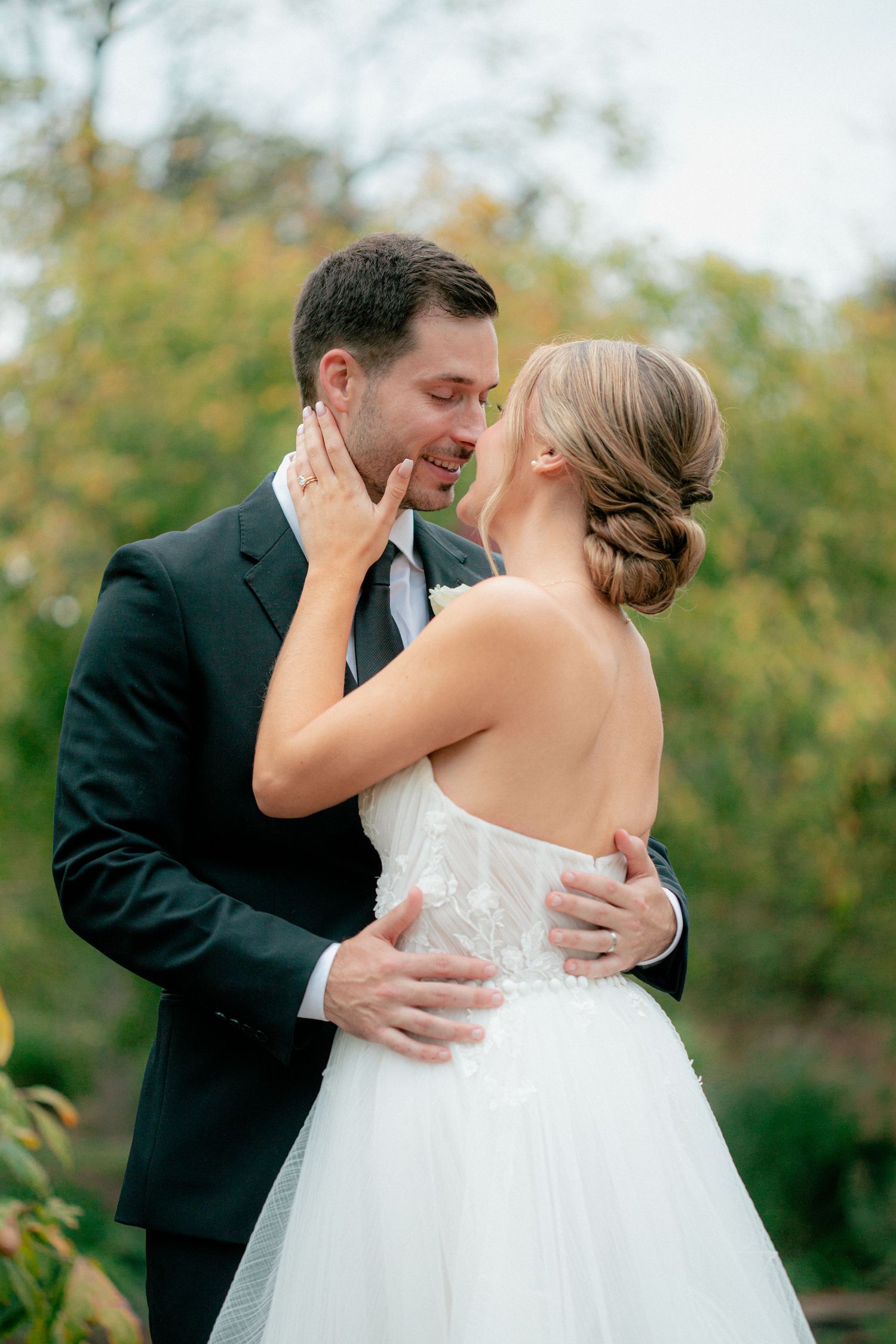 Bride and groom share an intimate almost-kiss embrace outdoors surrounded by lush autumn foliage.