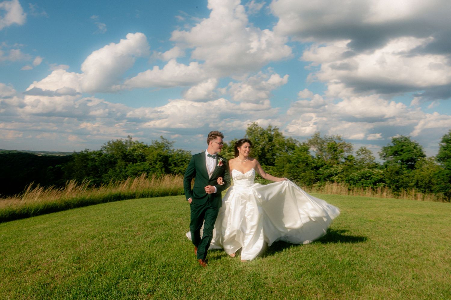 A bride and groom walk hand-in-hand across a lush green meadow under a bright blue sky with fluffy white clouds.