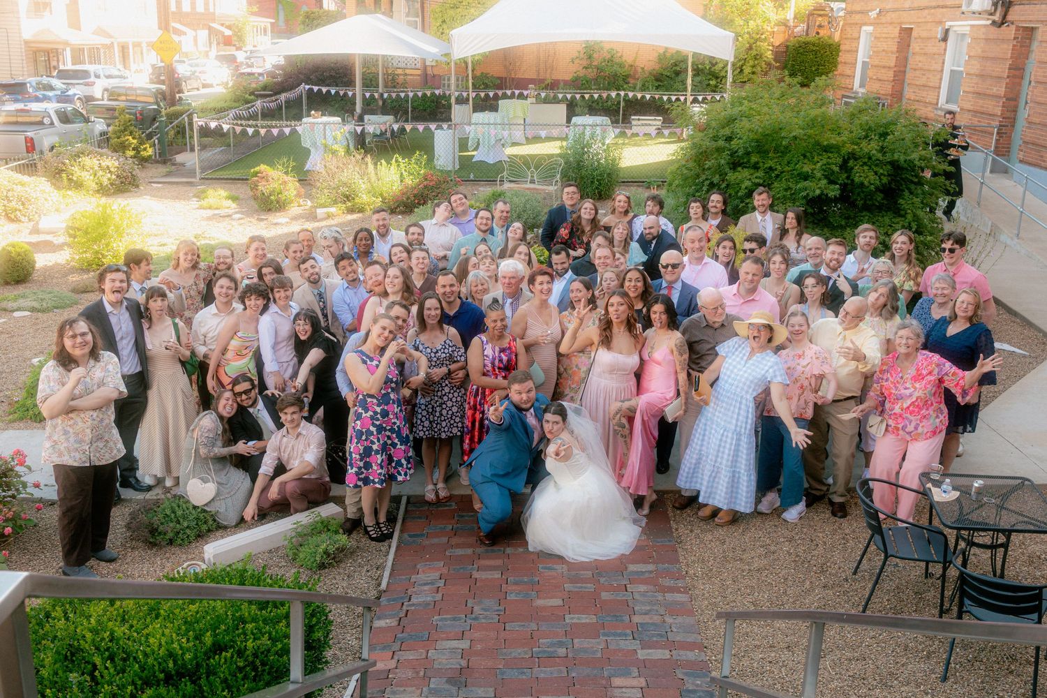 Large wedding group photo outdoors on brick patio with bride in white gown surrounded by guests in formal attire.