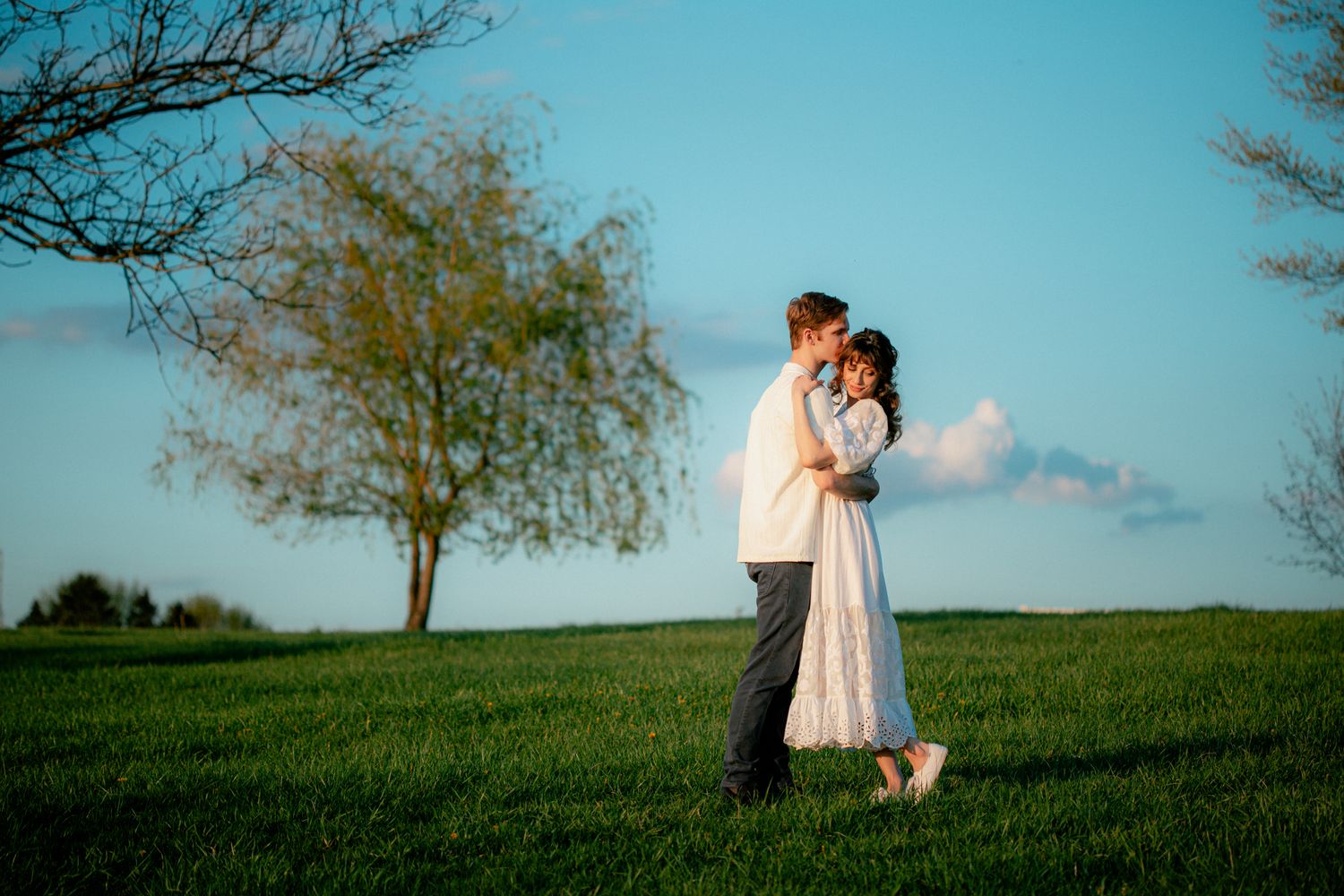 Couple sharing a romantic embrace in a lush green field under a blue sky with a lone tree.