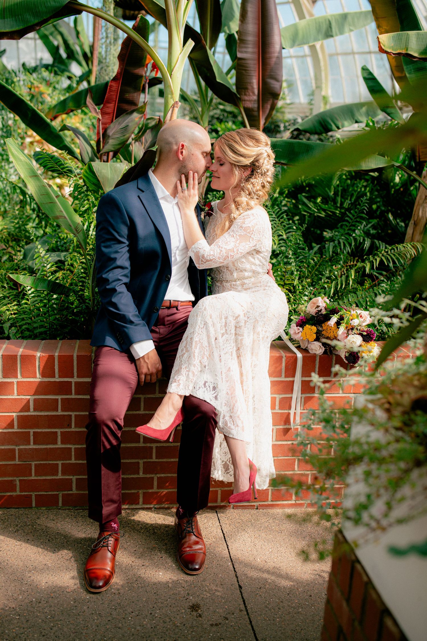 Bride and groom share a kiss while seated on a brick wall in a tropical greenhouse.