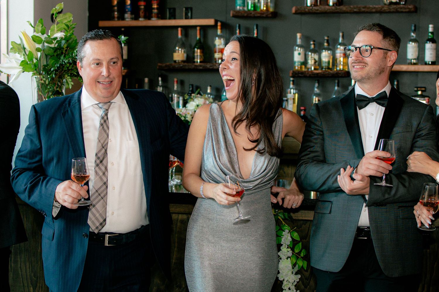Wedding guests laughing and holding drinks at bar during elegant reception celebration.