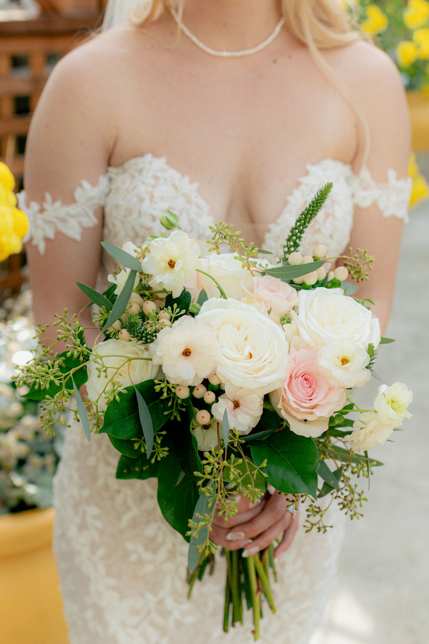 Bride in off-shoulder lace wedding gown holding a soft bouquet of white and blush pink roses.