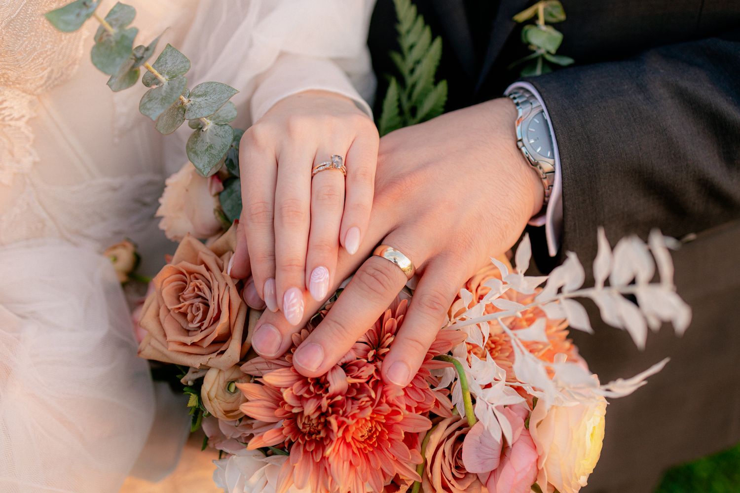 Bride and groom resting hands on a coral and peach bouquet, showing off their matching wedding bands.