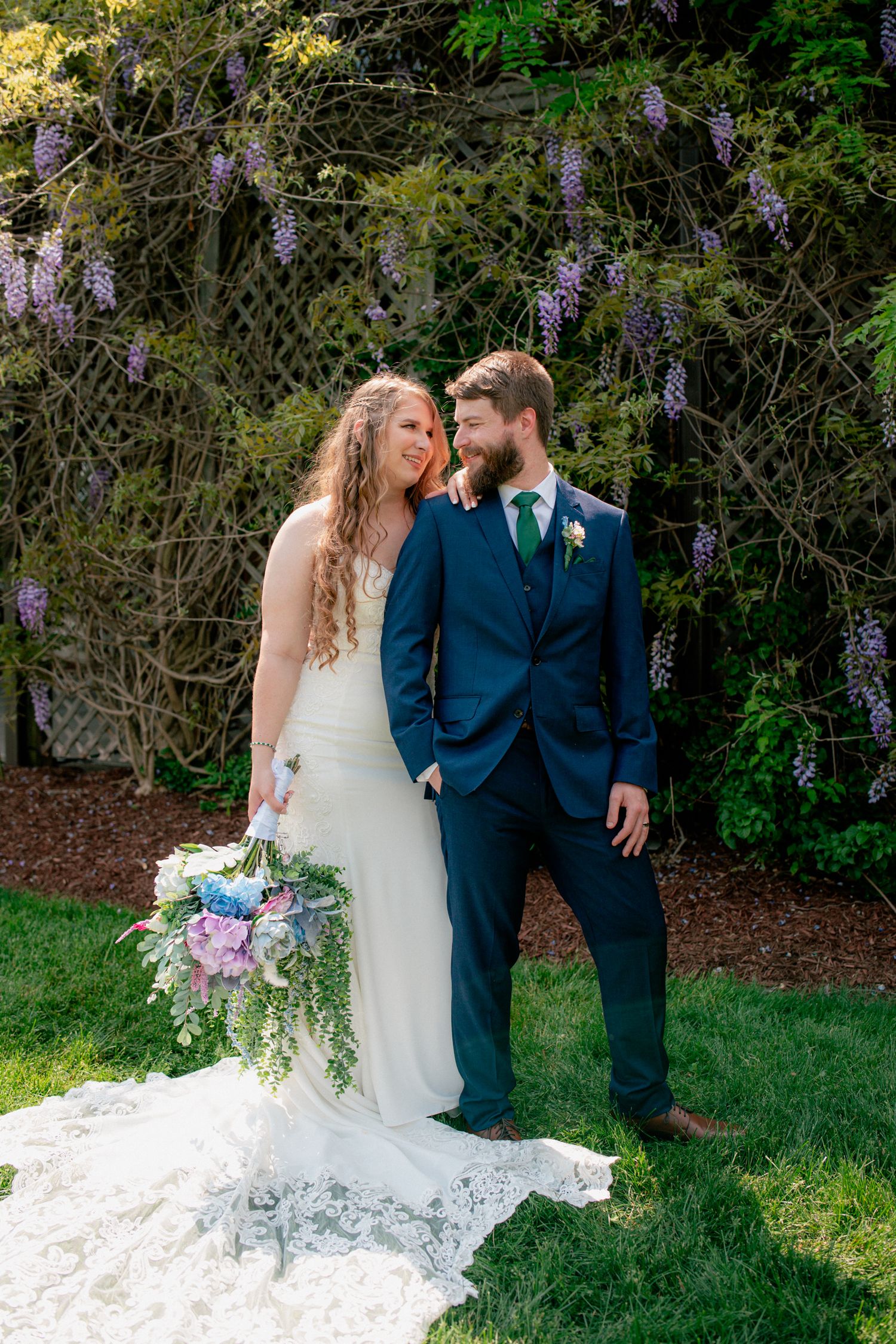 Smiling bride and groom pose under blooming purple wisteria, bride holding a lush wildflower bouquet.