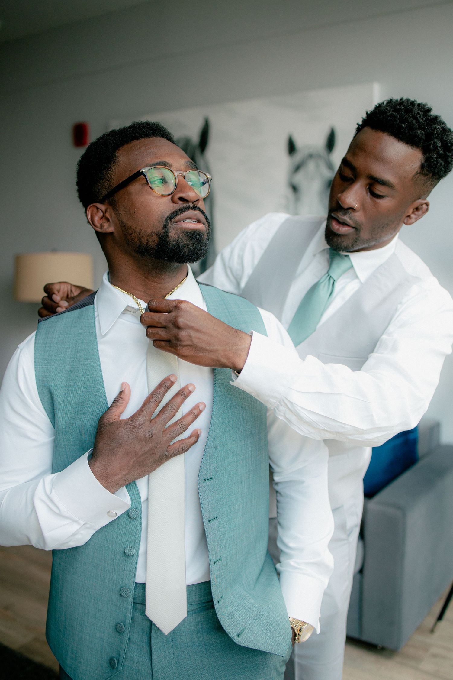 Groomsman in white suit adjusts tie of groom in teal vest and glasses before wedding.