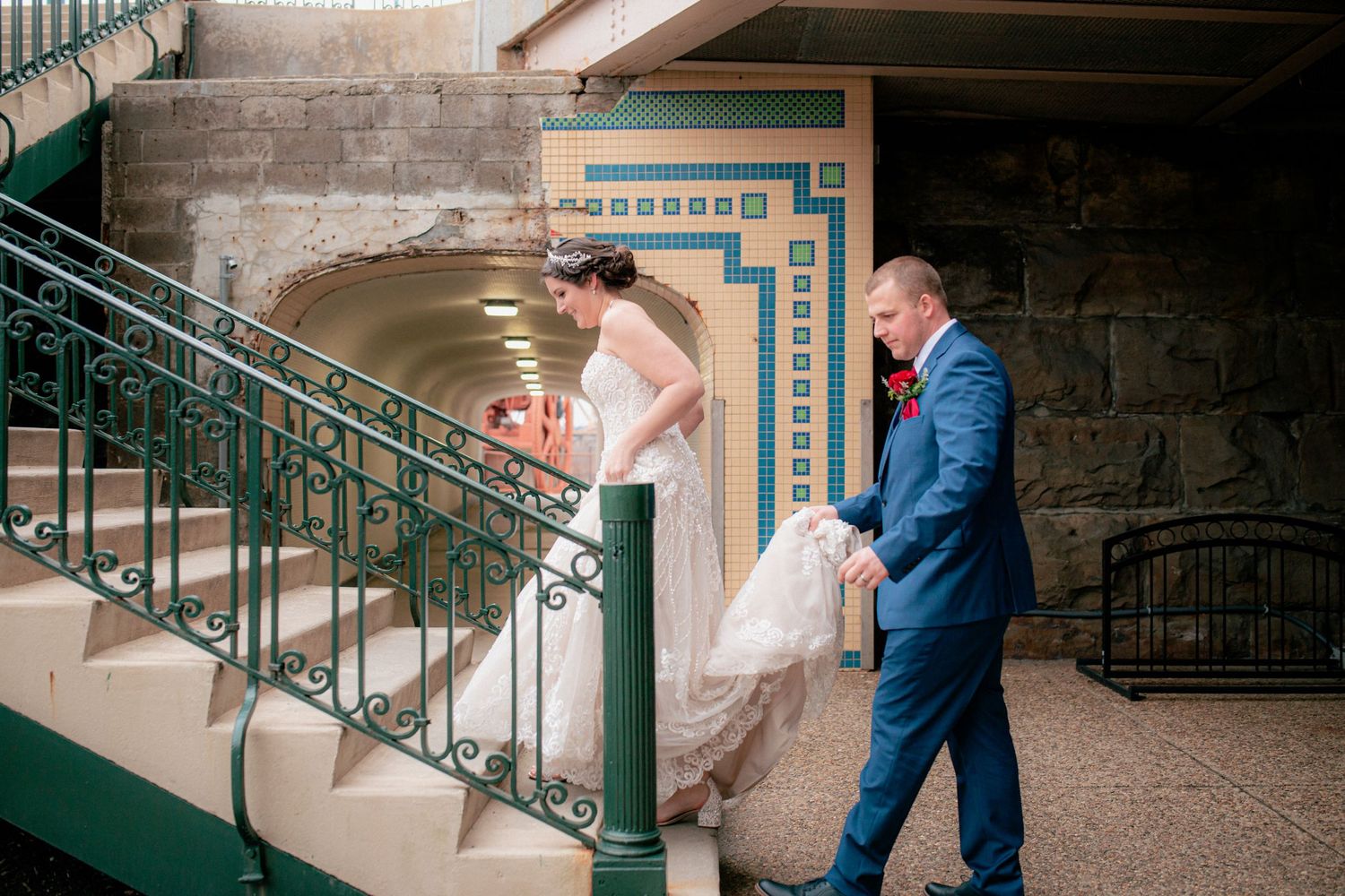 Groom in blue suit helps bride in lace gown up ornate green staircase with art deco details.