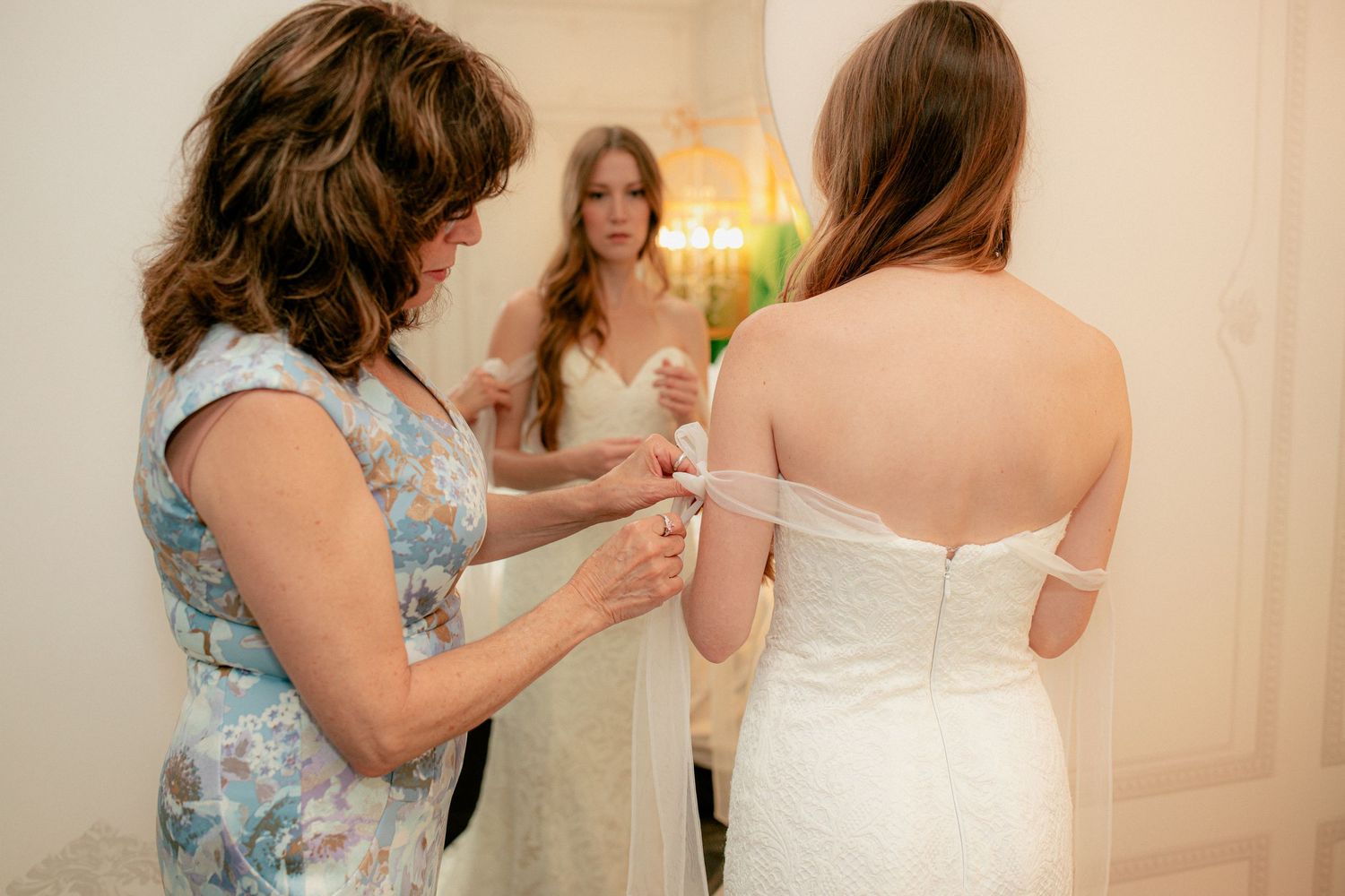 Mother helping bride button up wedding dress while reflection shows in mirror during bridal preparation.
