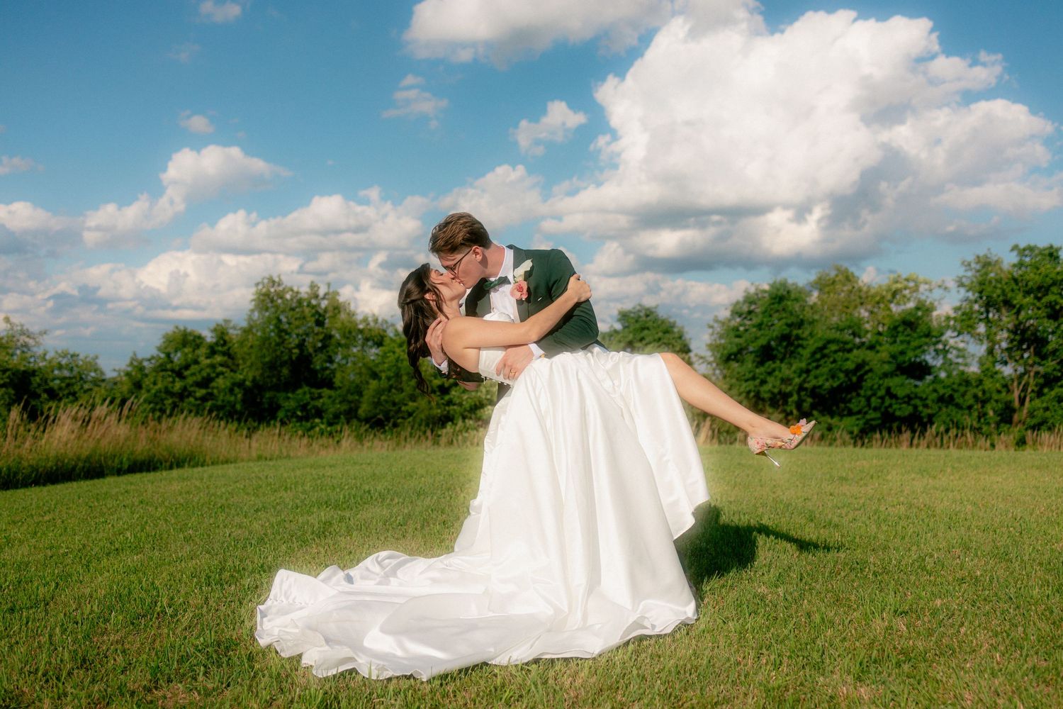 Groom dips bride in white gown for romantic kiss on green lawn under blue sky with fluffy clouds.