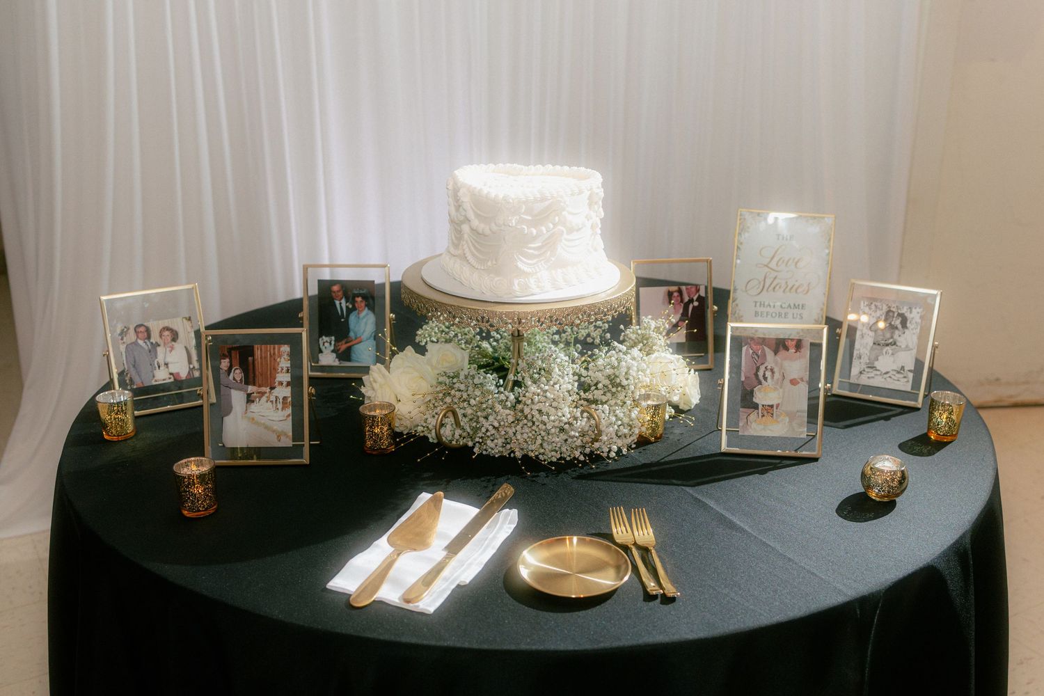 White wedding cake on black tablecloth surrounded by framed photos and floral arrangement with gold accents.
