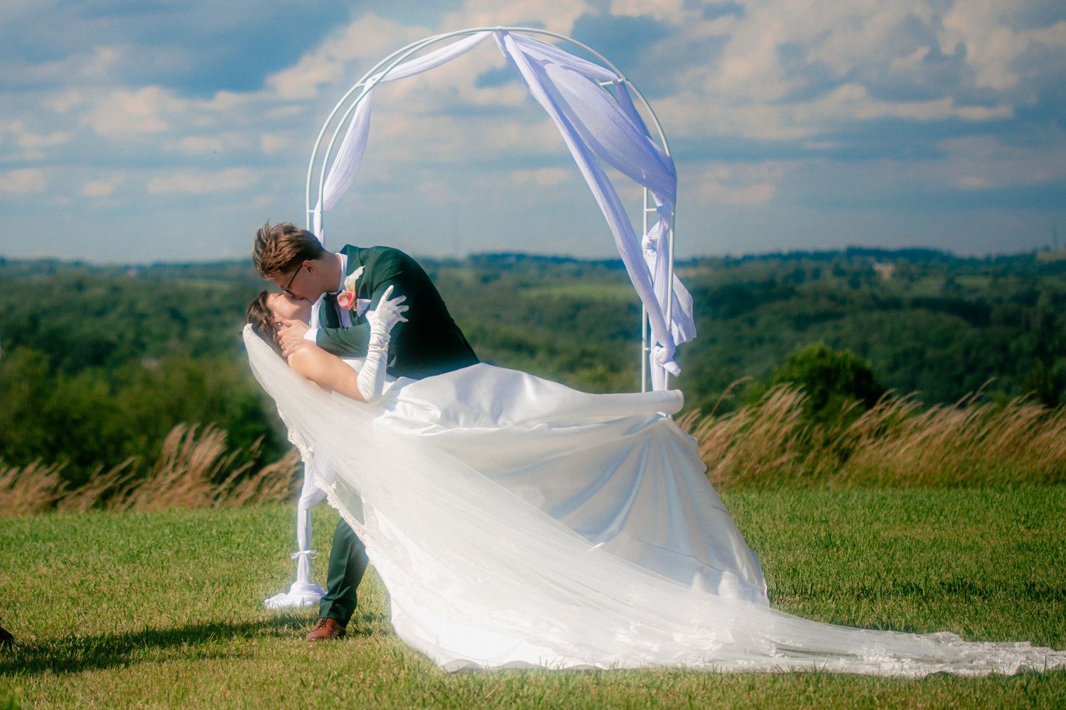 Bride and groom sharing a romantic dip kiss outdoors under a white arch on a green hillside on their wedding day.