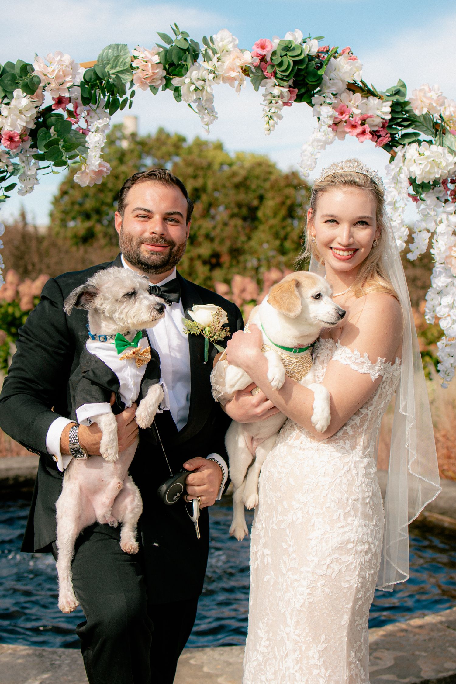 Smiling bride and groom holding their dogs under a floral arch at their outdoor wedding ceremony.