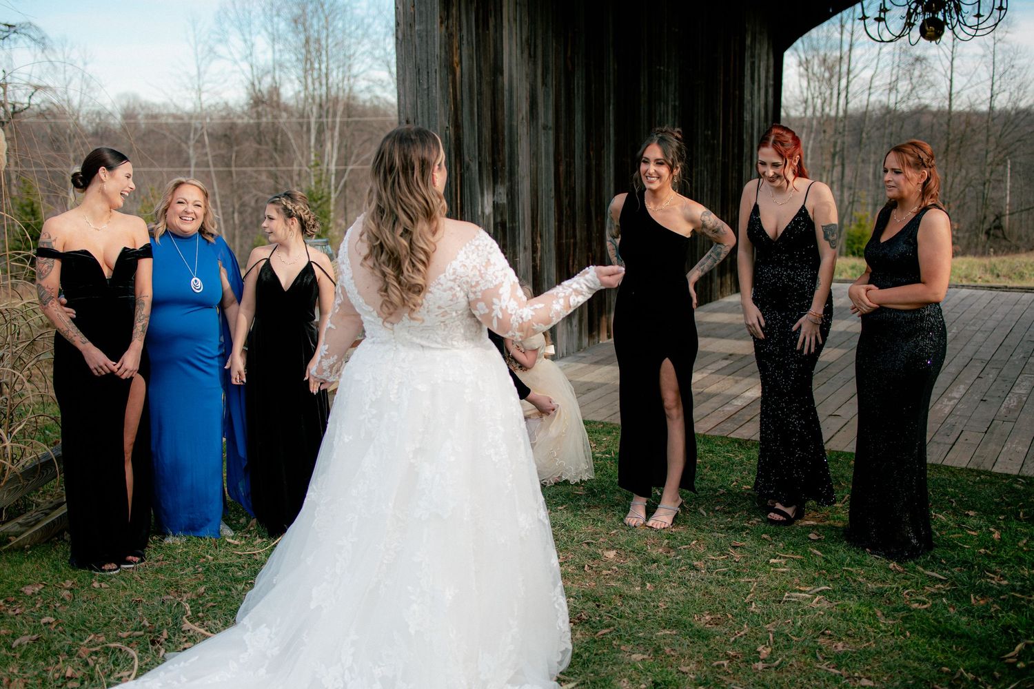 Bride reveals wedding dress to bridesmaids in black and blue gowns outside a rustic barn venue.