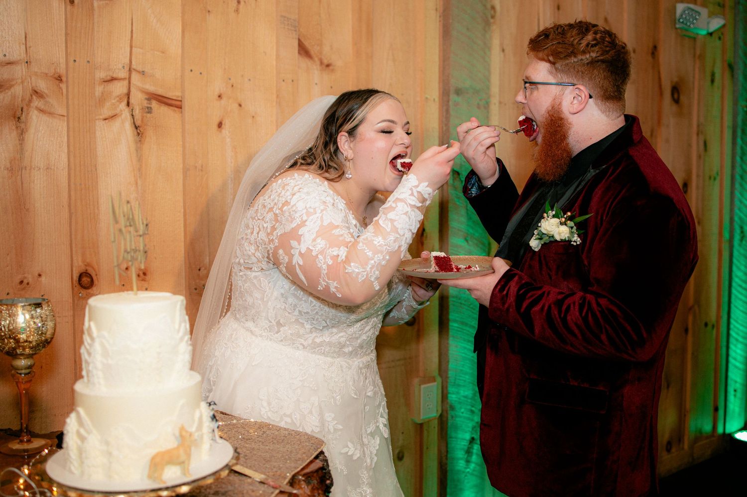 Bride and groom feeding each other wedding cake at rustic barn reception.