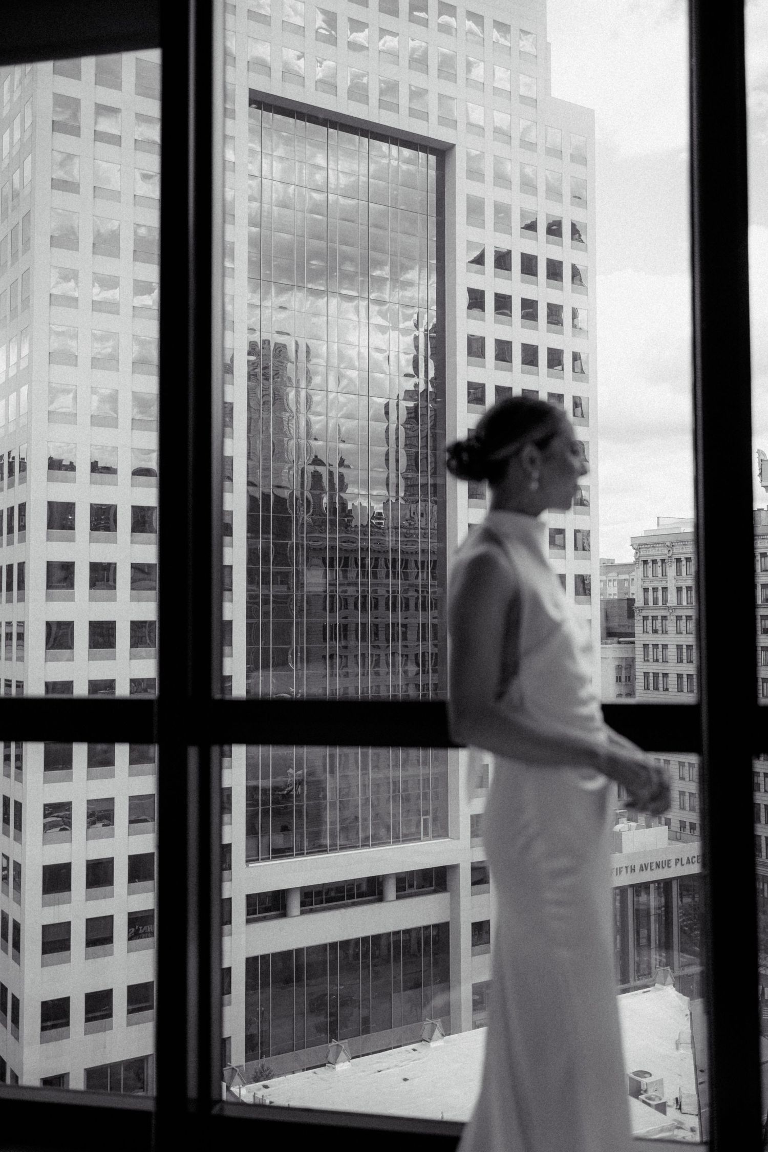 Black and white silhouette of bride in white gown standing before large window with city skyline.