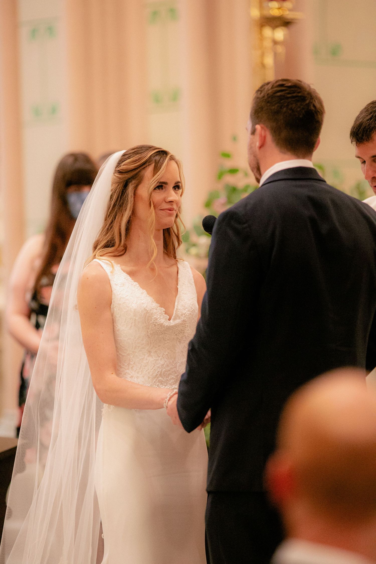 Radiant bride in white lace gown and veil holds hands with groom at church altar ceremony.