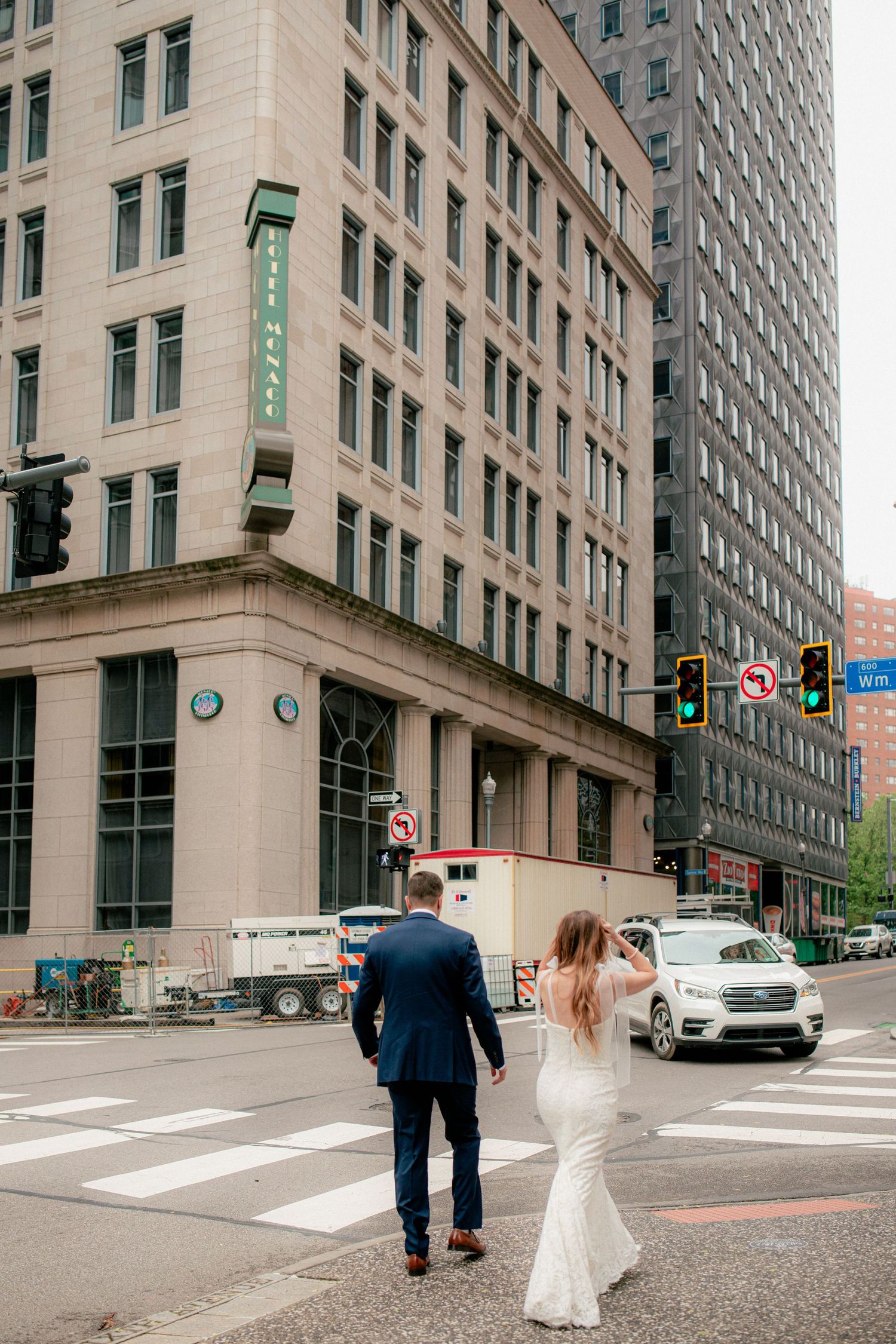 Bride and groom in wedding attire cross downtown city street intersection surrounded by tall buildings.