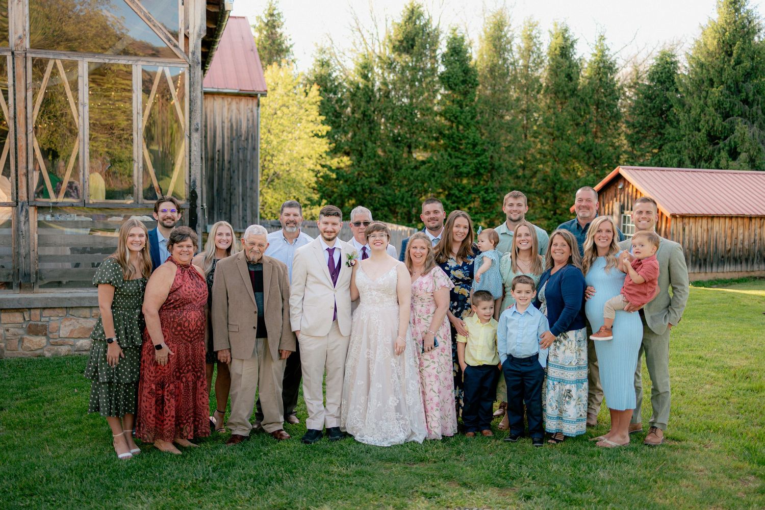 Large wedding party posing outdoors near a rustic barn, bride in white gown surrounded by family and friends on a sunny day.